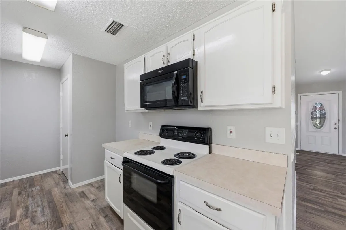 Kitchen features white cabinetry with a built-in microwave. A window and front door with glass inset are visible in the connected living space beyond. Wood-look vinyl flooring extends throughout, transitioning to the entry.