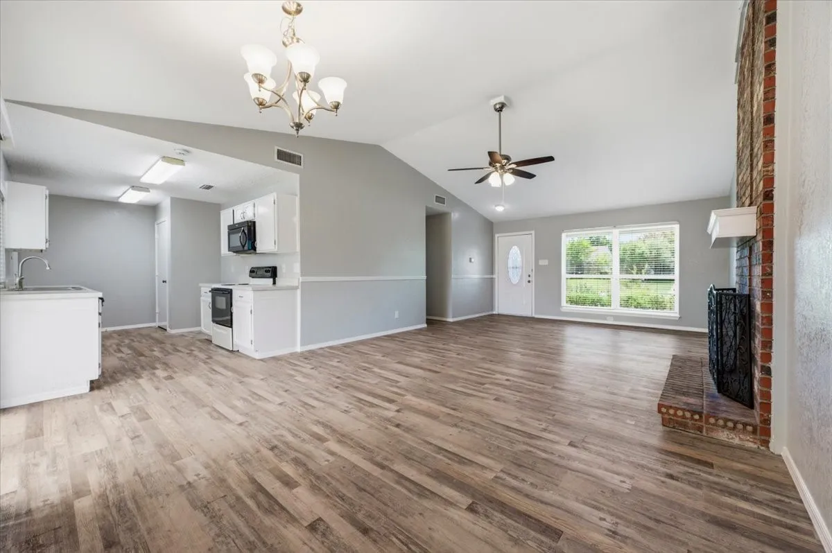 Living room features a vaulted ceiling with ceiling fan and light fixture, and a brick fireplace. A front door with glass inset and large window frame the space, which opens to the kitchen with white cabinetry and a built-in microwave. Wood-look vinyl flooring extends throughout.