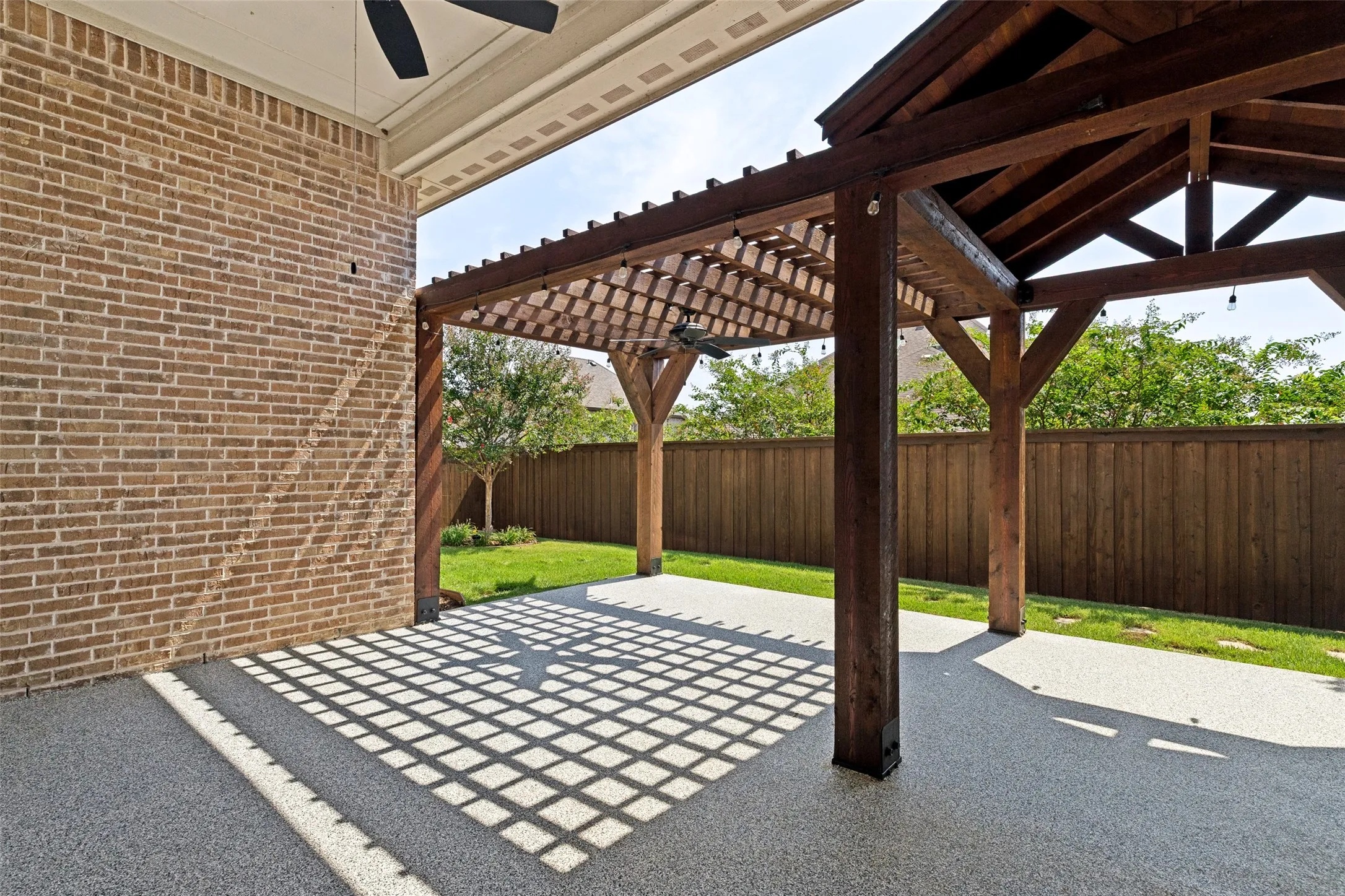 Covered patio with a wood pergola structure, textured concrete flooring, brick exterior wall, and a wooden privacy fence