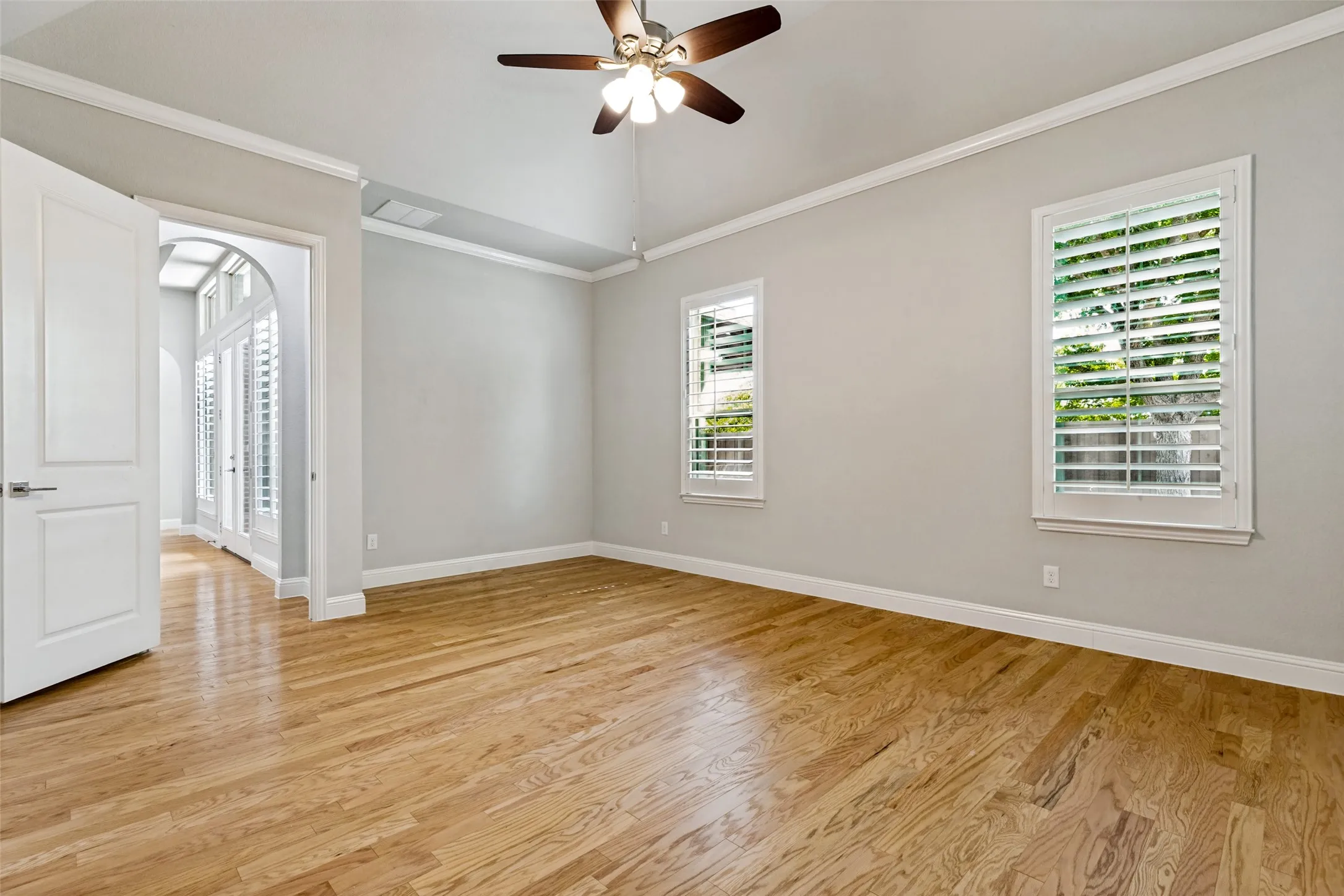 Vaulted ceiling with a ceiling fan, hardwood flooring, dual windows with plantation shutters, and an arched doorway