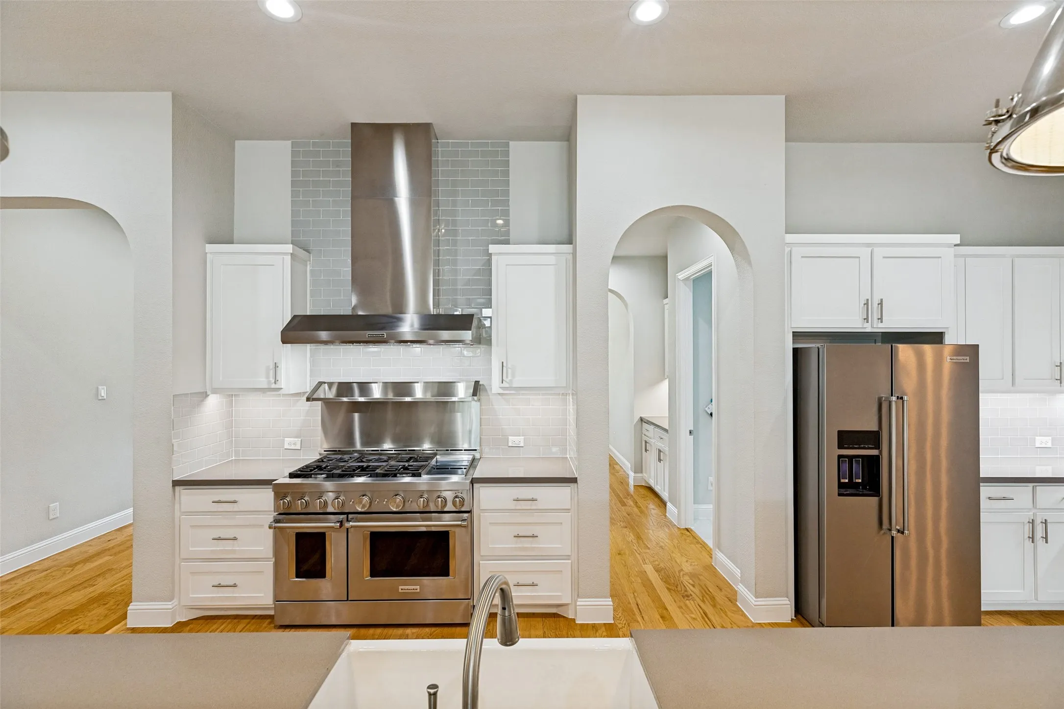 White cabinetry, stainless steel appliances, wood-finish flooring, recessed lighting, and an arched entryway