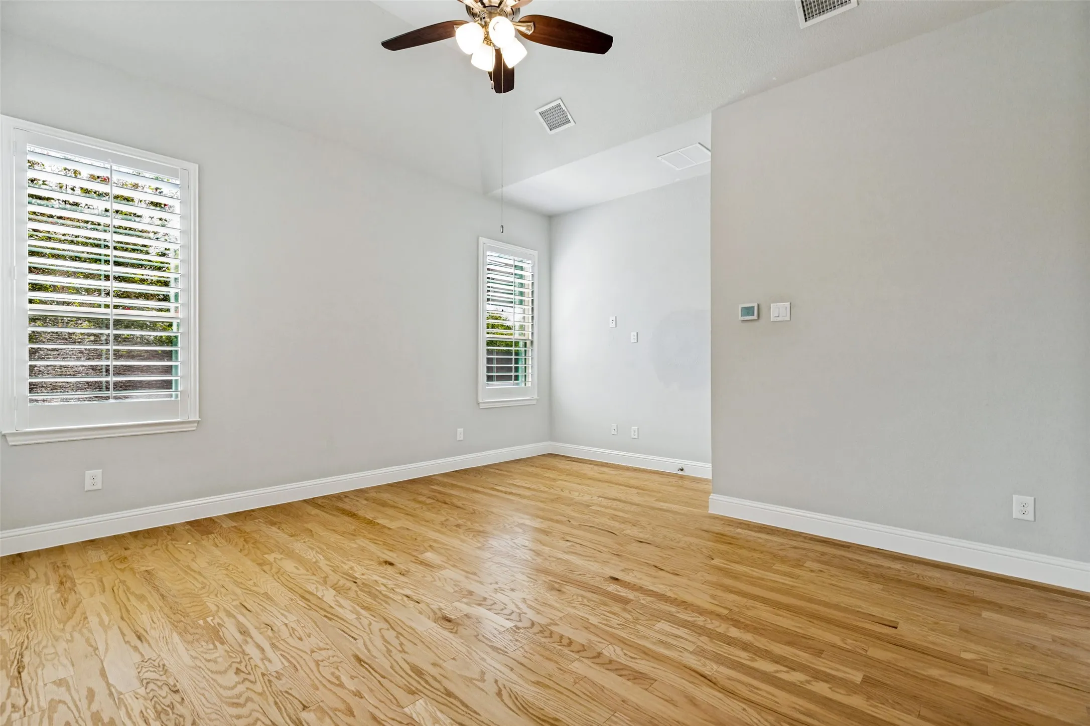 Wood-finish flooring, two windows with plantation shutters, ceiling fan with integrated lighting, and light gray wall paint