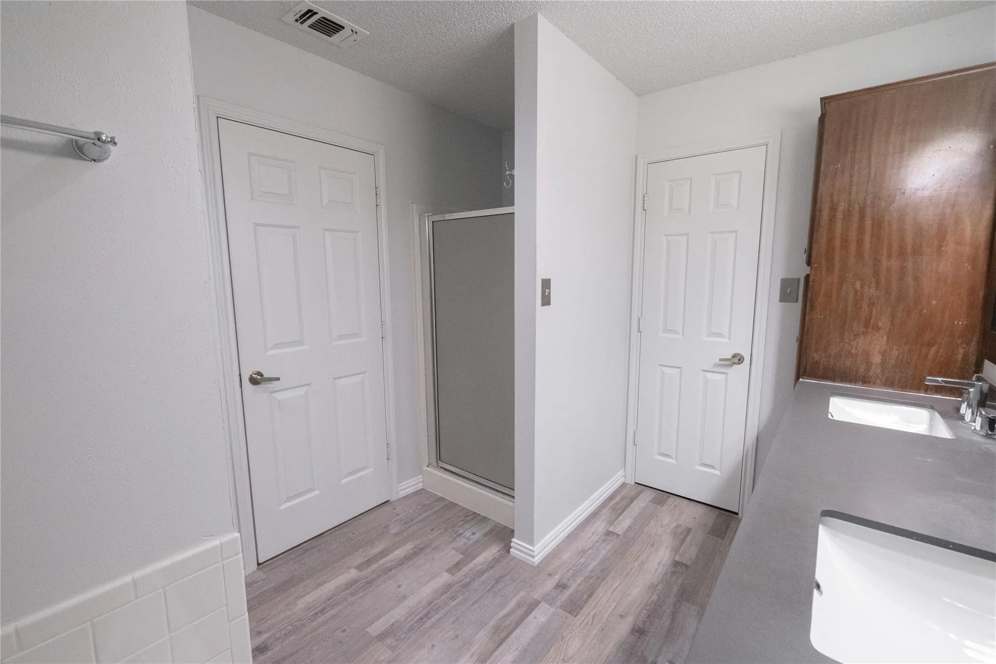Dual vanity with integrated sinks, wood-finish flooring, glass-enclosed shower, white paneled doors, and a wood-finish cabinet