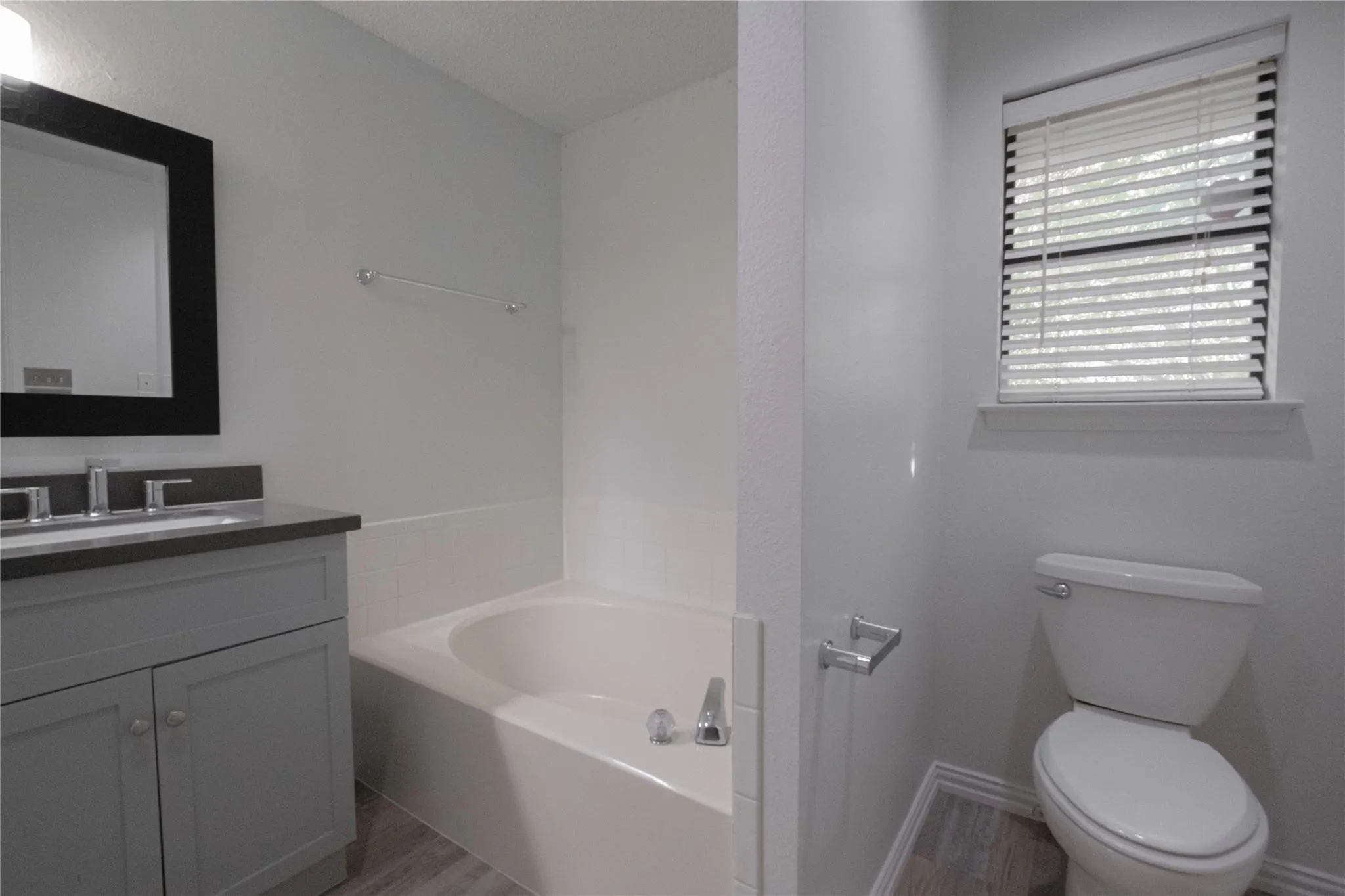 Vanity with dark countertop, large framed mirror, built-in bathtub, and wood-finish flooring