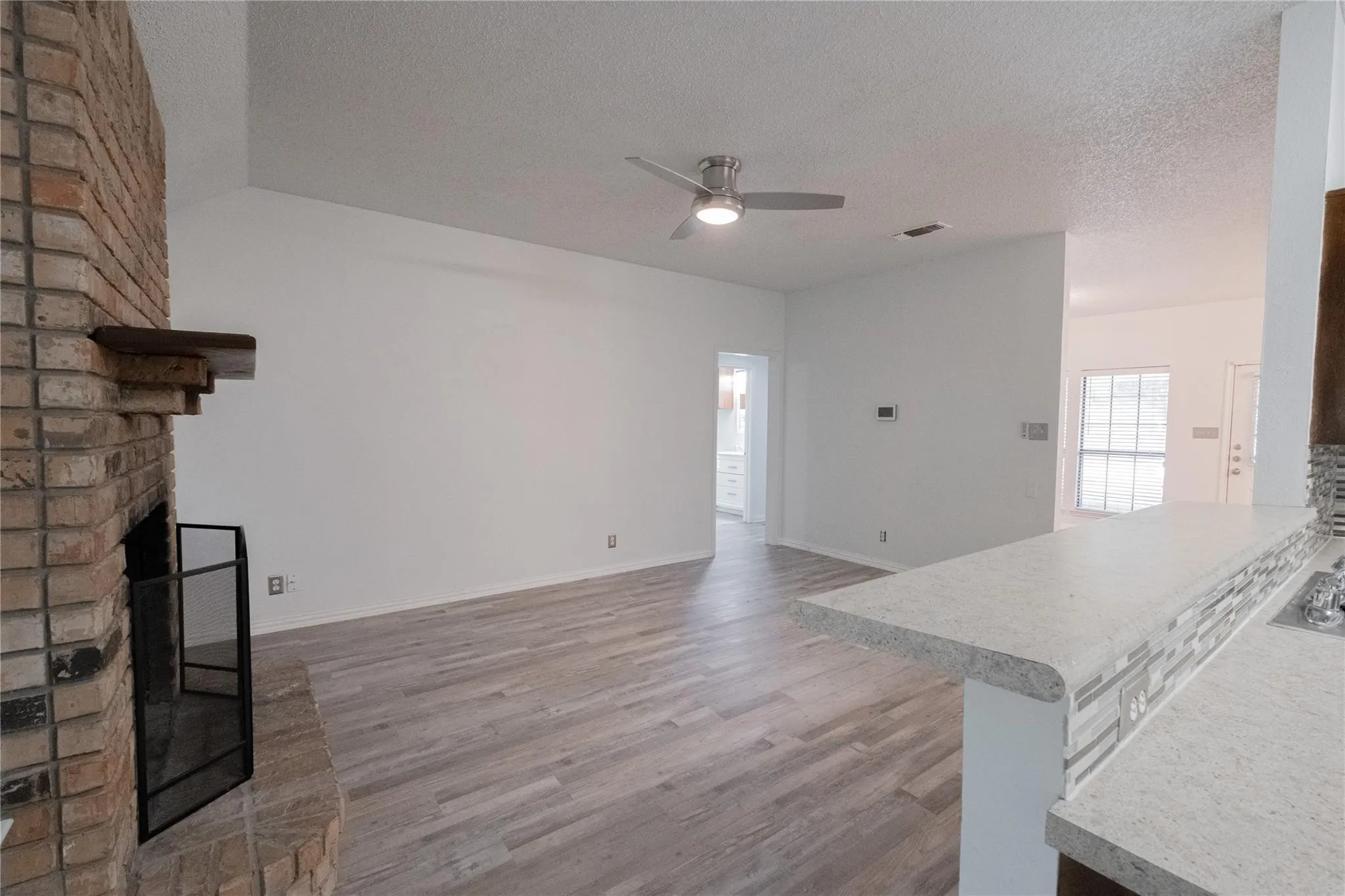 Exposed brick fireplace with a wood mantle, wood-finish flooring, ceiling fan with integrated lighting, and a breakfast bar with stone-finish countertops and a mosaic tile backsplash