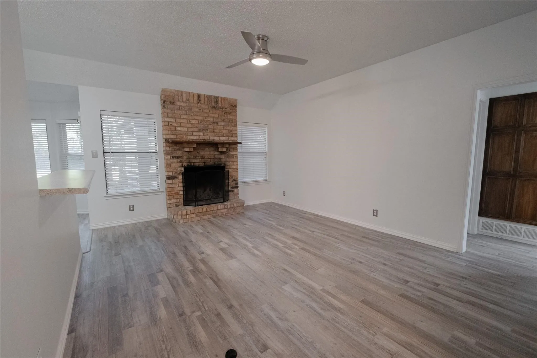Spacious living area featuring light wood-finish flooring, a brick fireplace with a wood mantle, and a ceiling fan with integrated lighting