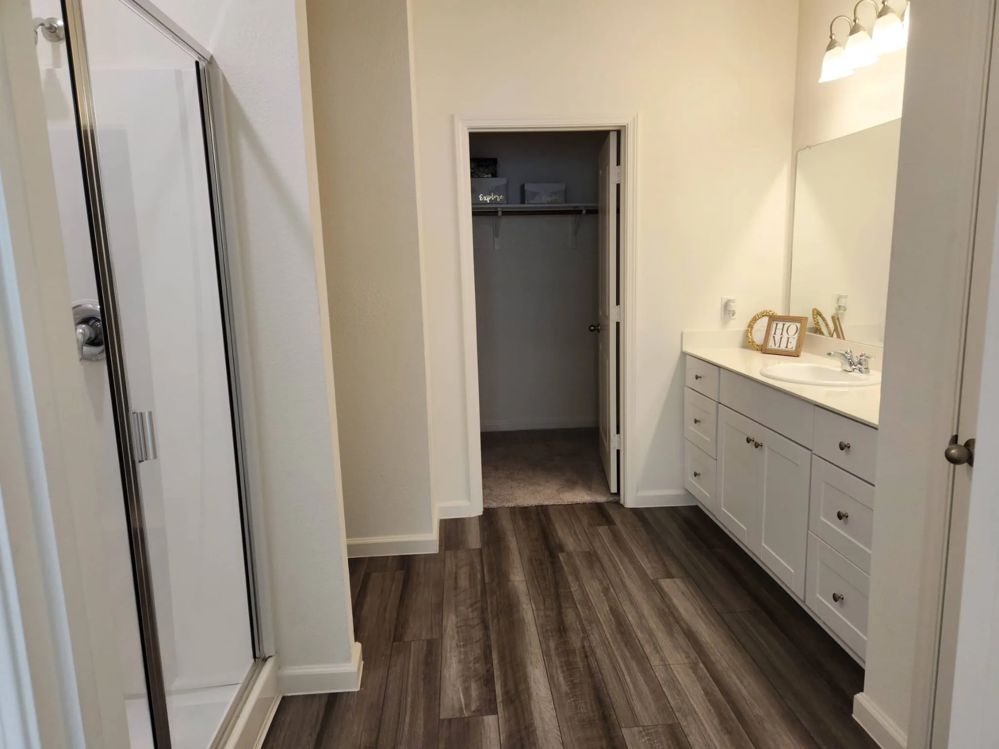 Walk-in shower with sliding glass door, wood-finish flooring, white vanity with integrated sink, and wall-mounted vanity lighting