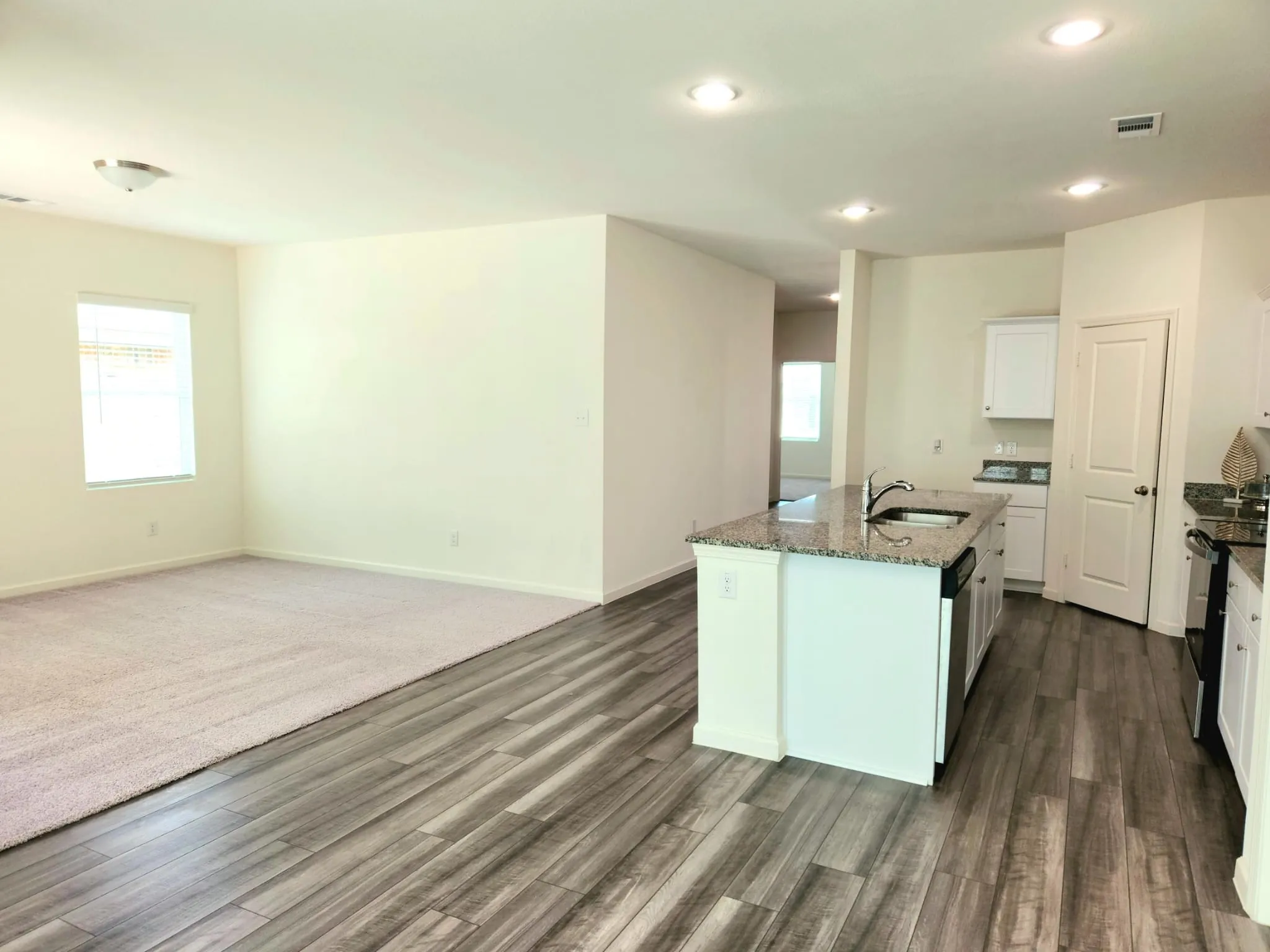 Open-concept living area featuring wood-finish flooring, recessed lighting, a kitchen island with a sink, and white cabinetry with granite countertops