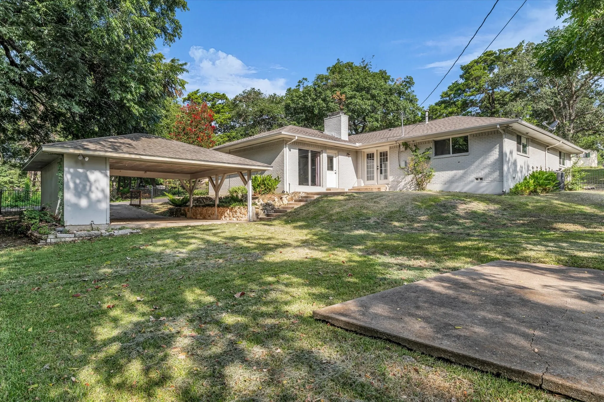 Exterior featuring a covered carport, mature trees, a concrete patio, and a sloped lawn