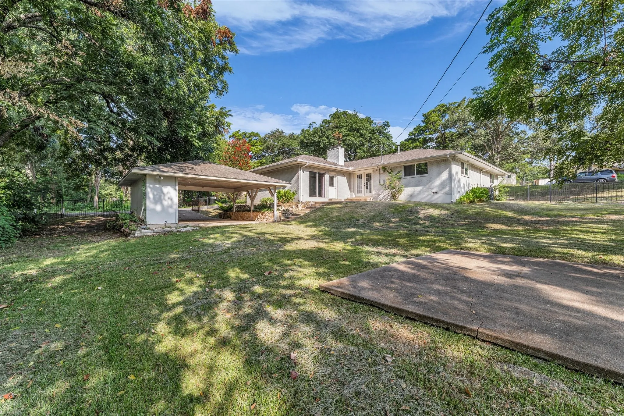 Expansive green space with a concrete slab, mature trees, and a covered carport structure