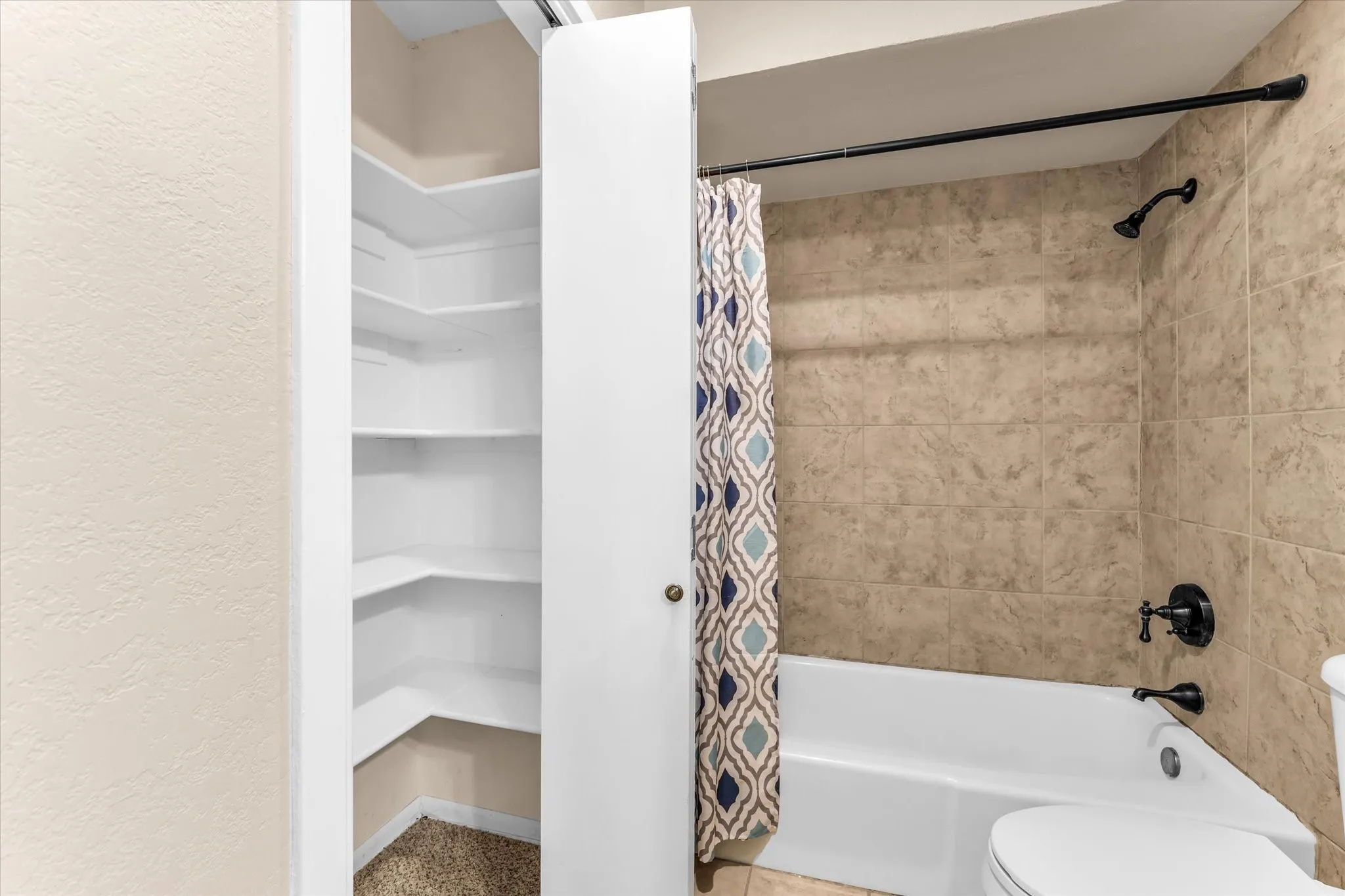Built-in shelving inside linen closet, tile shower surround, bathtub, and black-finish bath fixtures