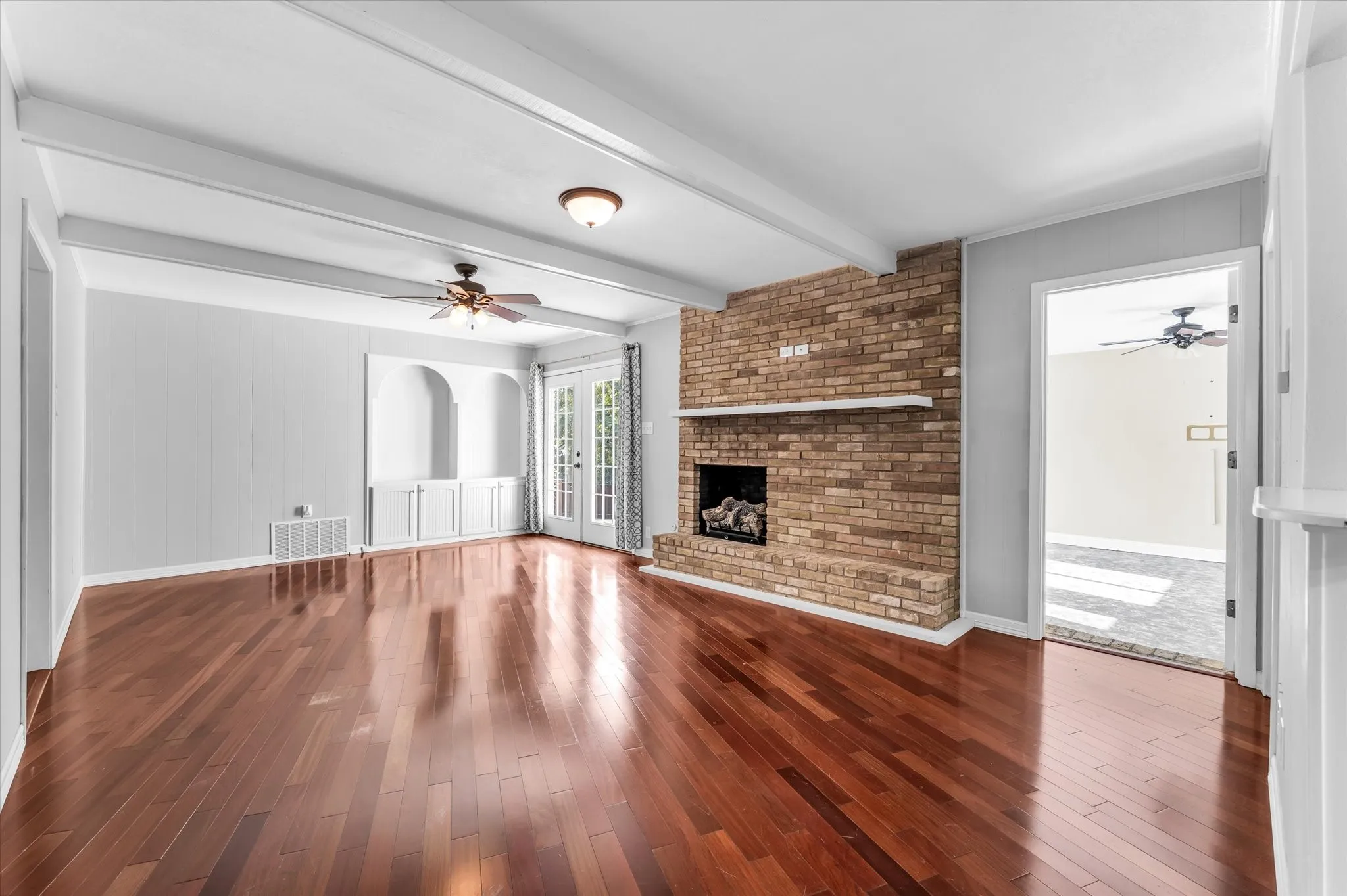 Hardwood flooring, exposed ceiling beams, brick fireplace with mantel, built-in cabinetry with arched niches, and French doors
