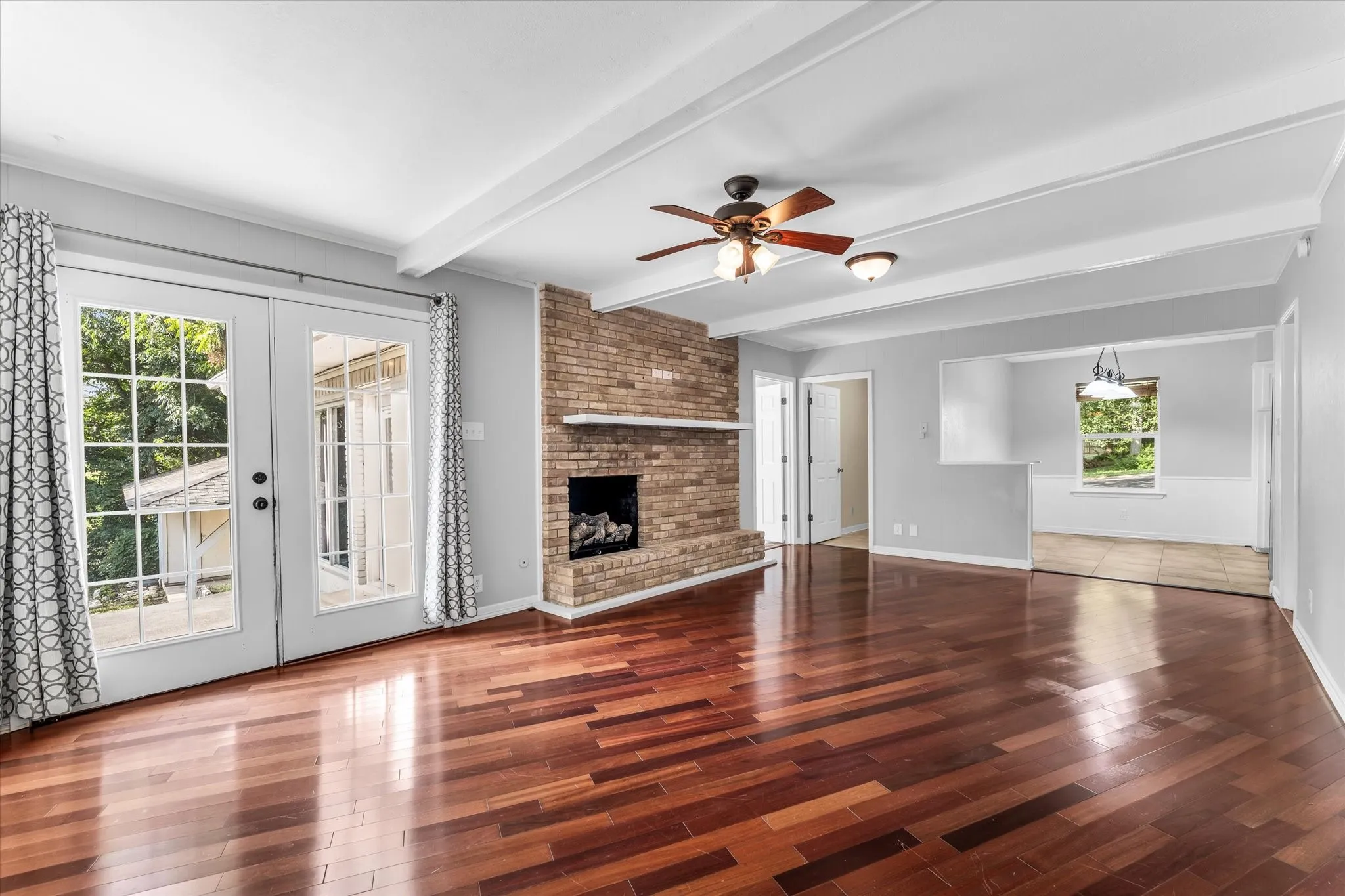 Wood-finish flooring, exposed ceiling beams, brick fireplace with mantel (shared with Den), French doors, and ceiling fan with integrated lighting