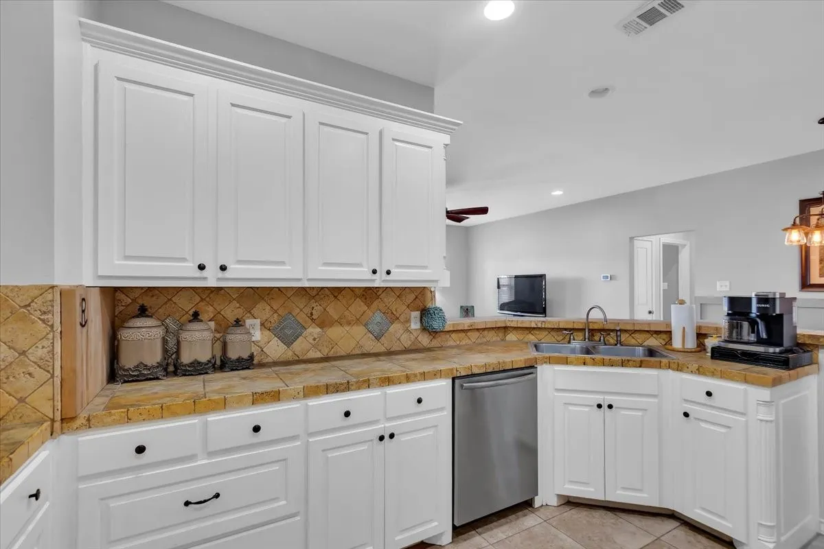 White shaker-style cabinetry with crown molding, natural stone backsplash with accent tiles, stainless steel dishwasher, and a dual-basin sink with a chrome faucet