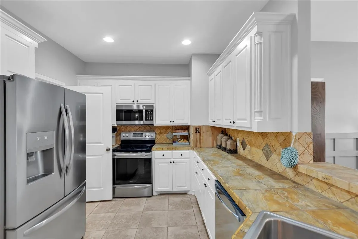 White cabinetry with crown molding, stainless steel appliances, tile flooring, stone-finish countertops, and a full-height tile backsplash