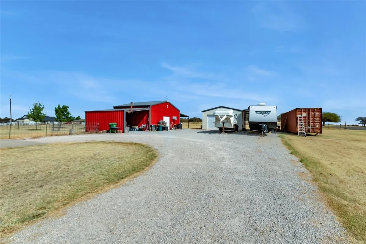 Gravel driveway accessing a red metal-sided barn with an attached open-sided structure, and a separate white metal-sided shed