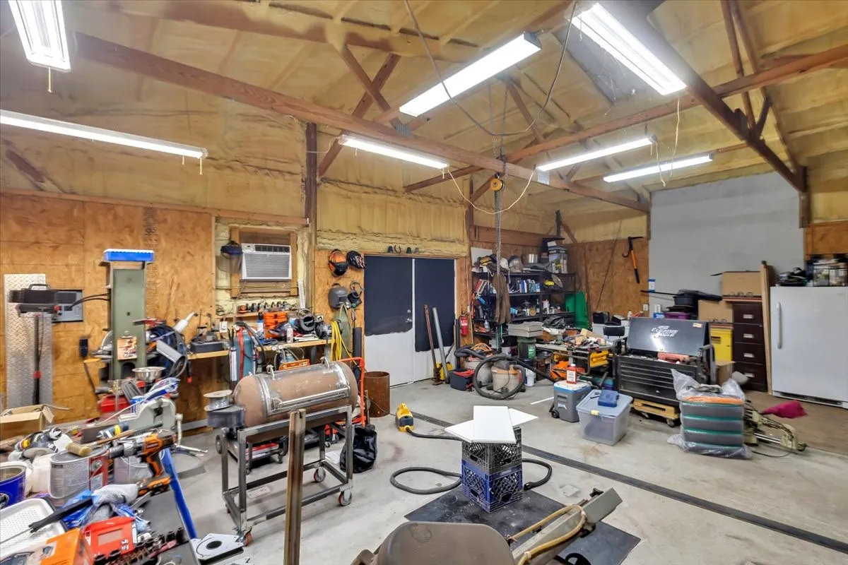 Spacious workshop with exposed wood beams, finished concrete flooring, multiple fluorescent light fixtures, and a wall-mounted air conditioning unit