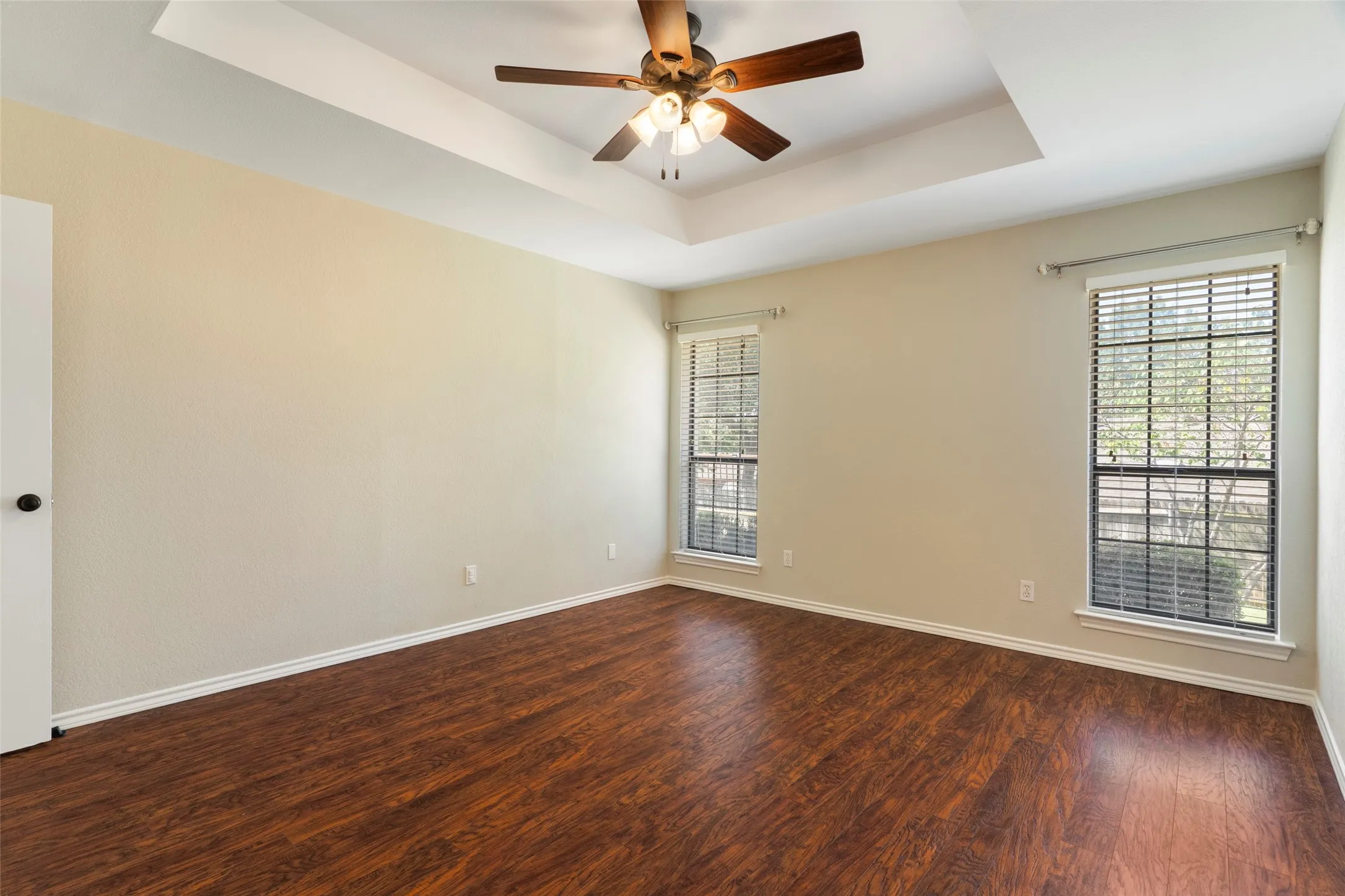 Tray ceiling with a ceiling fan, wood-finish flooring, and two windows with blinds
