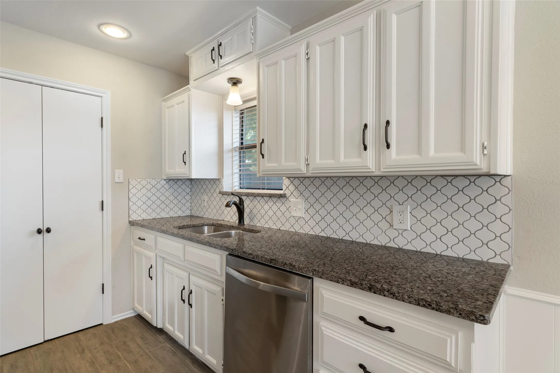 White shaker-style cabinetry, dark granite-style countertops, stainless steel sink, geometric tile backsplash, and wood-finish flooring