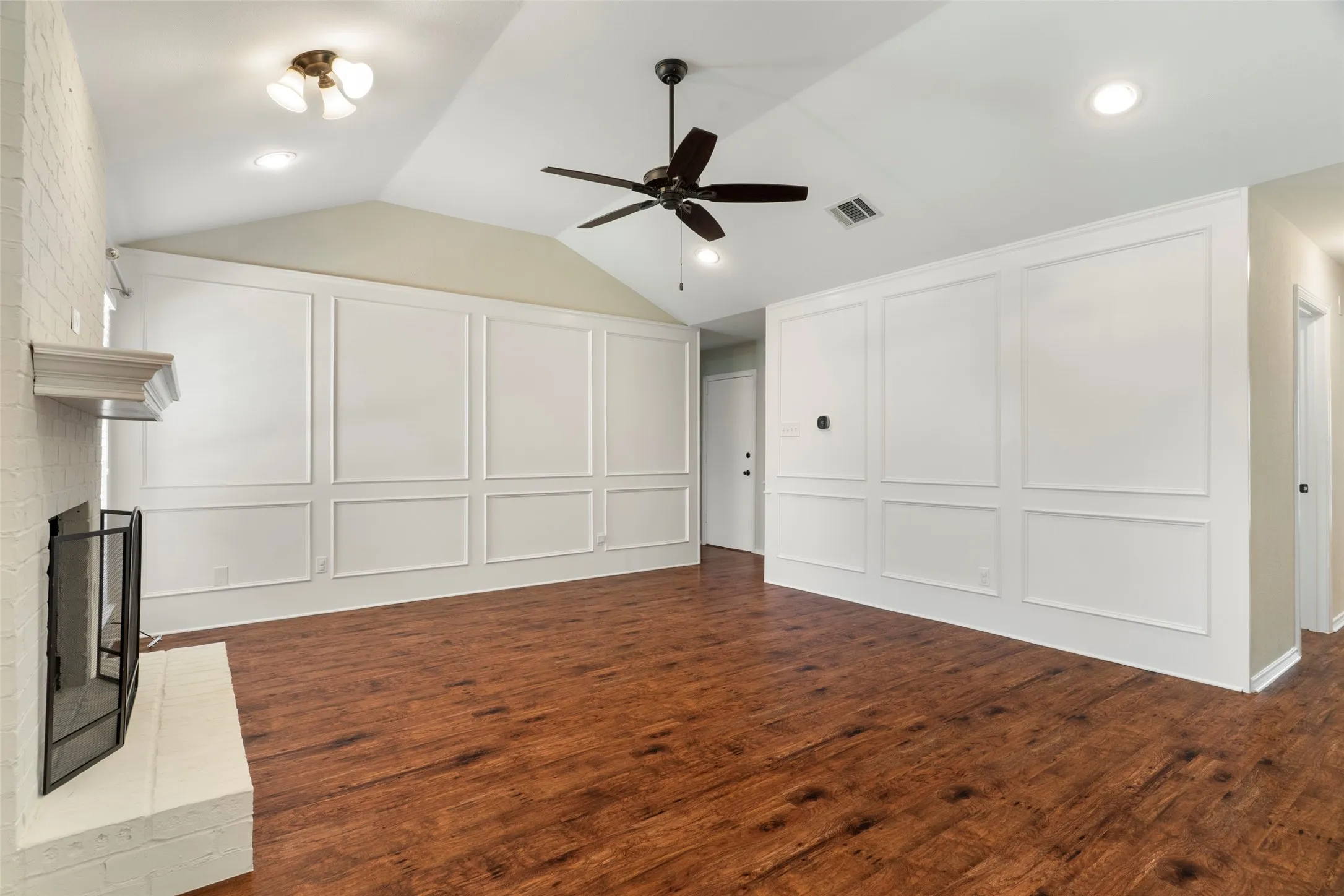 Vaulted ceiling with a ceiling fan, recessed lighting, white panel accent walls, wood-finish flooring, and a brick fireplace