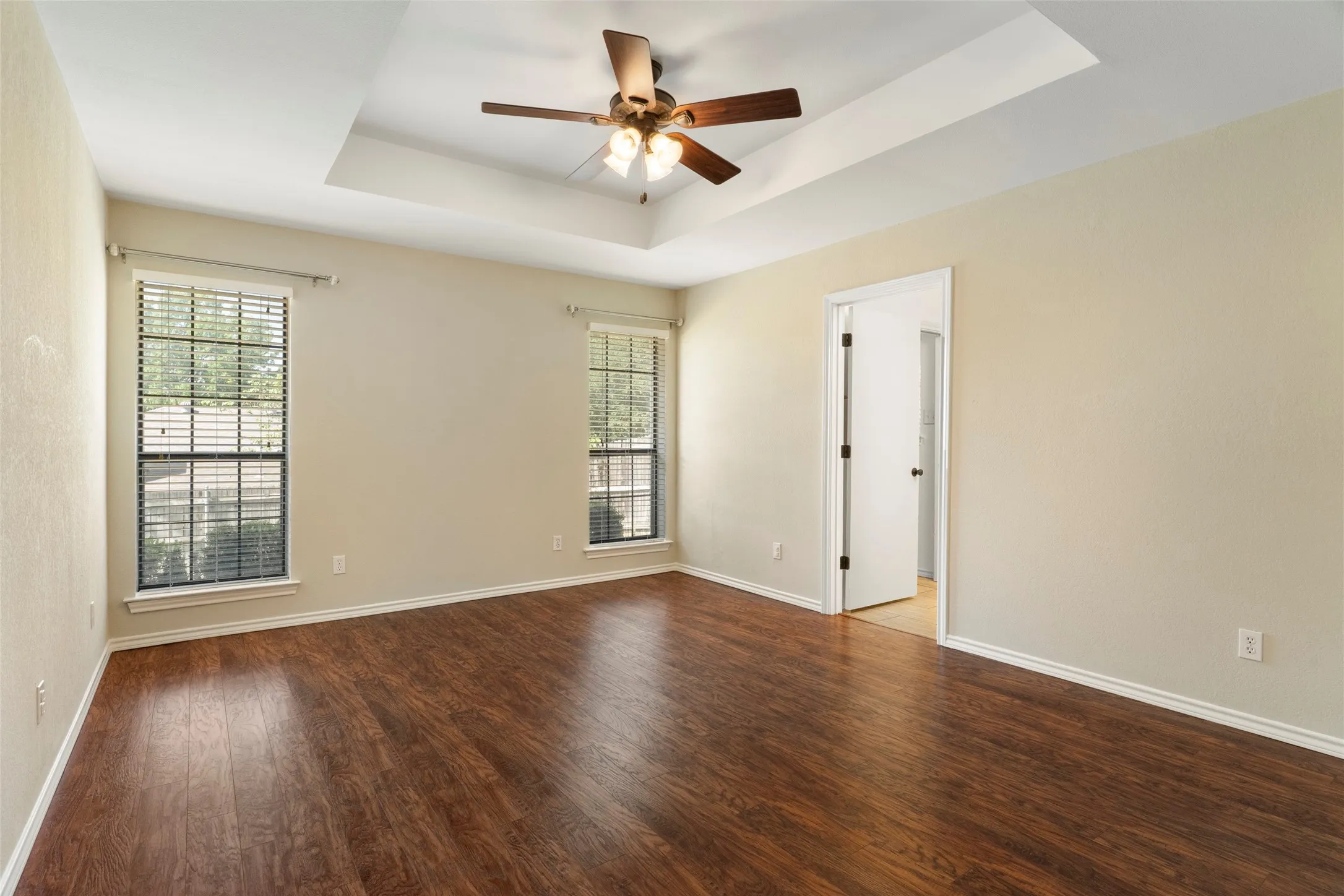 Tray ceiling with a built-in ceiling fan, wood-finish flooring, and two windows with blinds