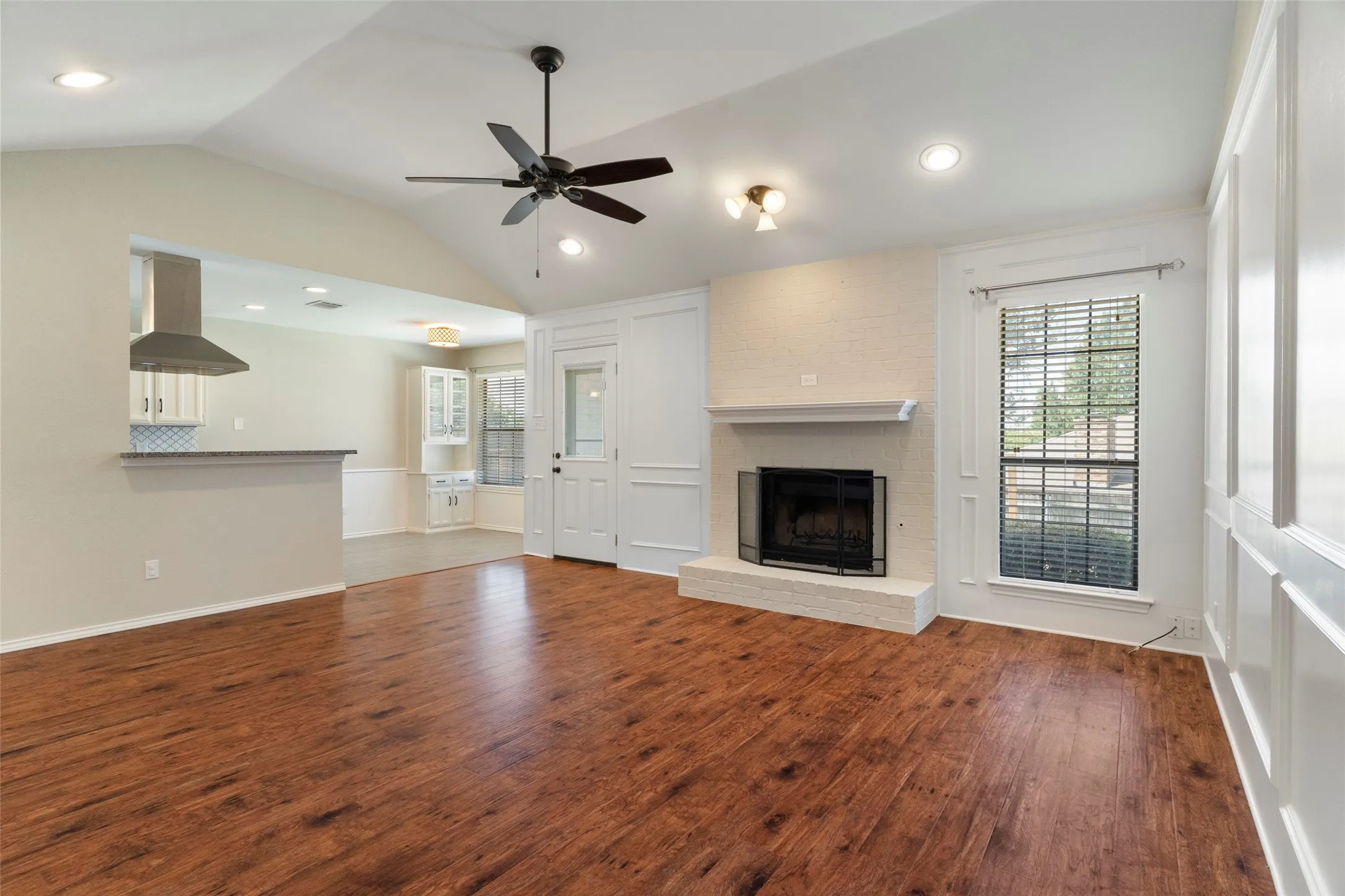 Vaulted ceiling with recessed lighting, wood-finish flooring, fireplace with white brick hearth, white paneling, and an expansive pass-through window