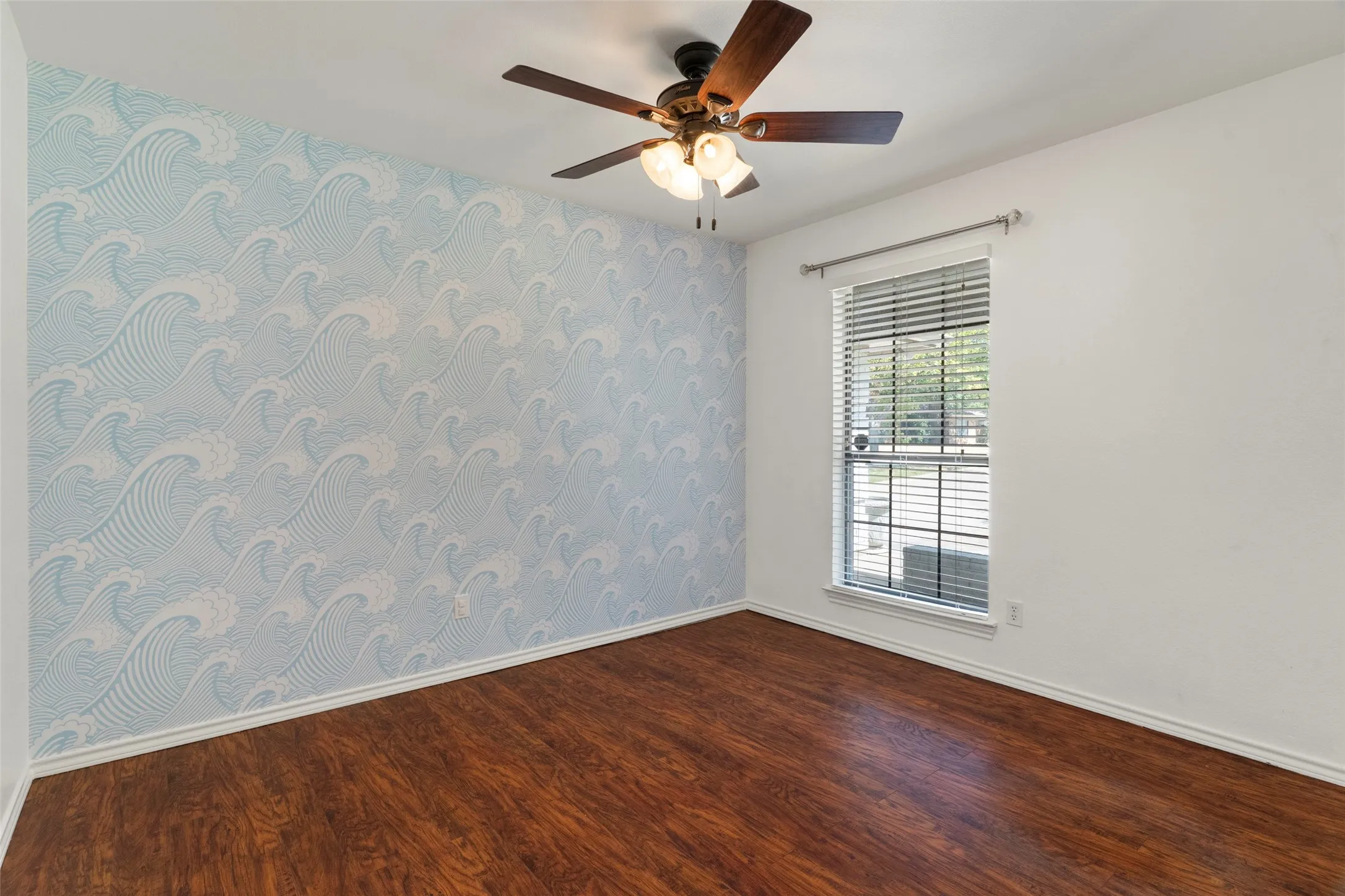 Wood-finish flooring, ceiling fan with integrated lighting, window with blinds, and decorative wallpaper accent wall