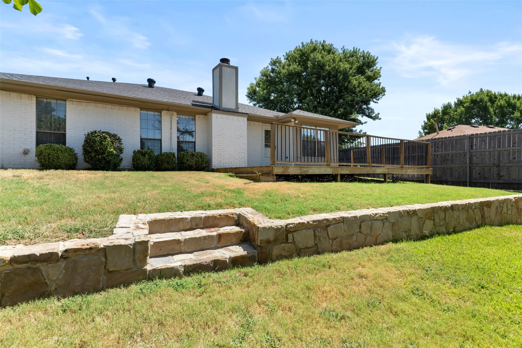 White brick exterior with multiple windows, elevated wood deck, and a stone retaining wall featuring stone steps