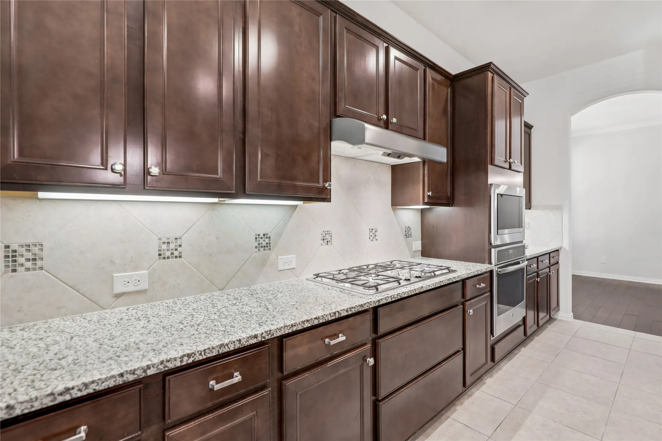 Rich wood cabinetry with brushed nickel hardware, granite countertops, tile backsplash with mosaic accents, stainless steel built-in oven and microwave, and a gas cooktop