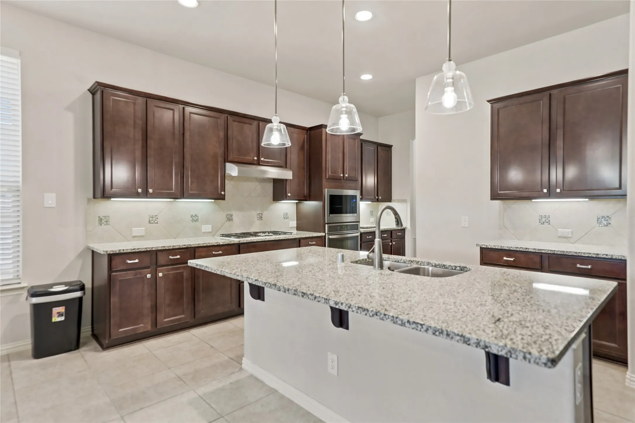 Kitchen featuring a central island with a double basin sink, granite countertops, dark wood-finish cabinetry, and a tile backsplash with decorative accents