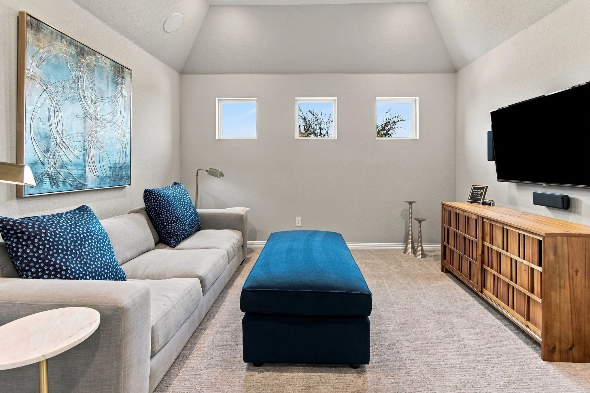 Vaulted ceiling, three square windows, built-in ceiling speaker, wall-mounted television, and warm wood-tone media console