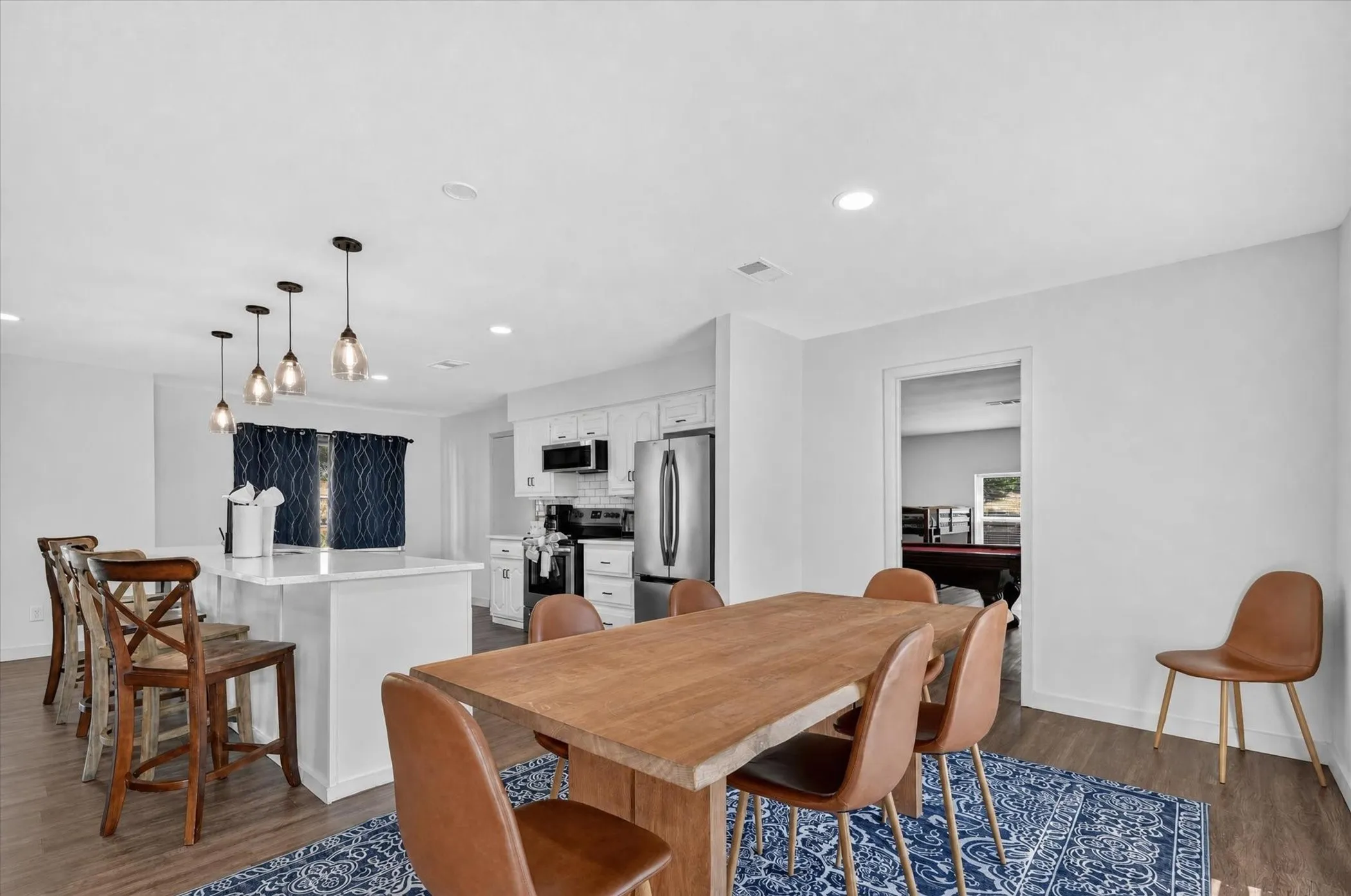 Open-concept dining and kitchen area featuring a wood-finish dining table, cognac-toned upholstered chairs, stainless steel appliances, white cabinetry, pendant lighting, and dark wood-finish flooring throughout