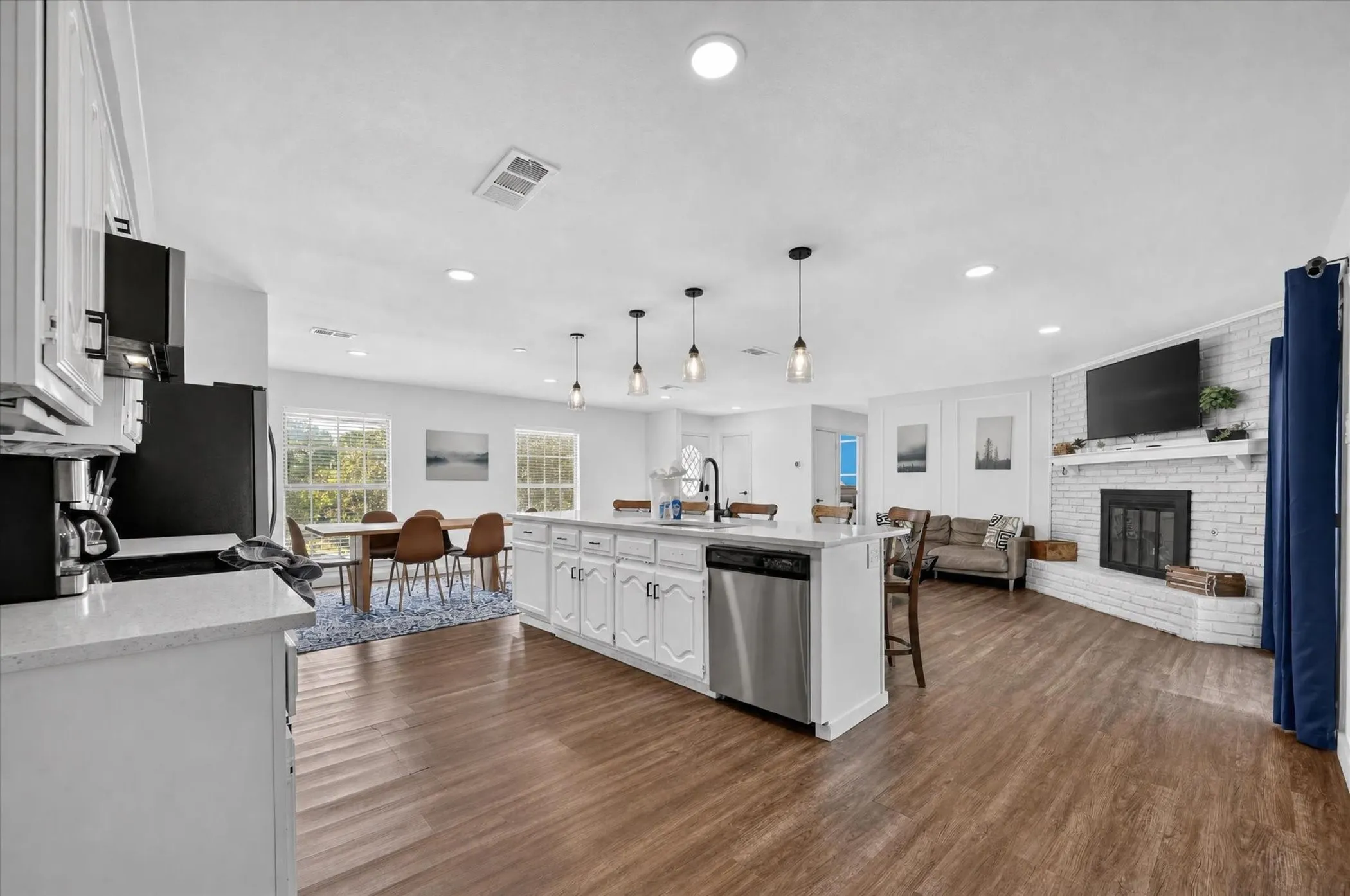 Wood-finish flooring throughout, white shaker-style cabinetry, a large kitchen island with pendant lighting, a white brick fireplace, and recessed ceiling lighting
