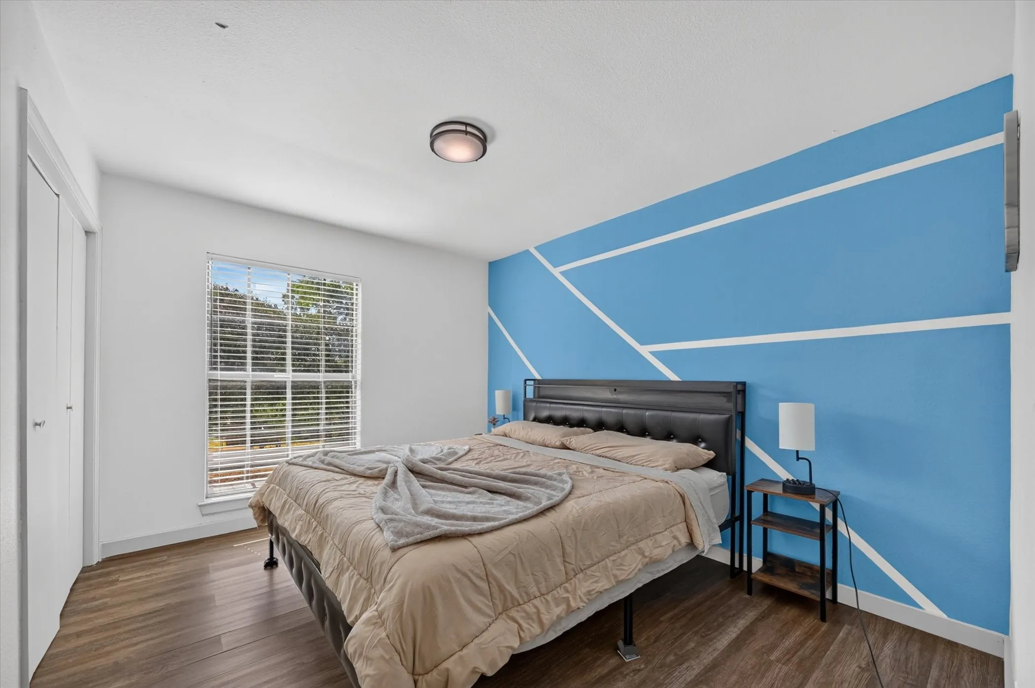 Wood-finish flooring, a window with horizontal blinds, and a flush-mount ceiling light fixture
