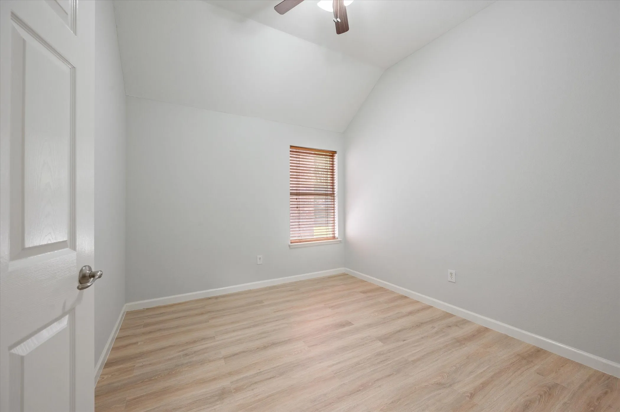 Vaulted ceiling, wood-finish flooring, single window with blinds, ceiling fan with integrated lighting, and white interior door with brushed nickel hardware