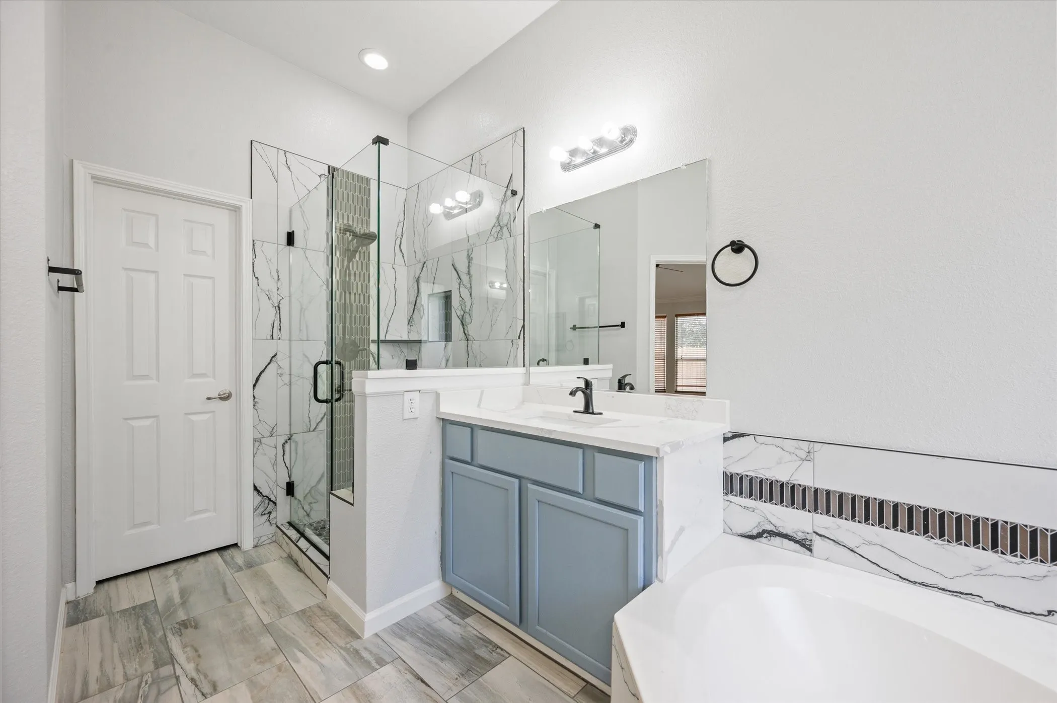 Tiled flooring, glass-enclosed shower, built-in soaking tub, vanity with light blue cabinetry, and wall-mounted mirror
