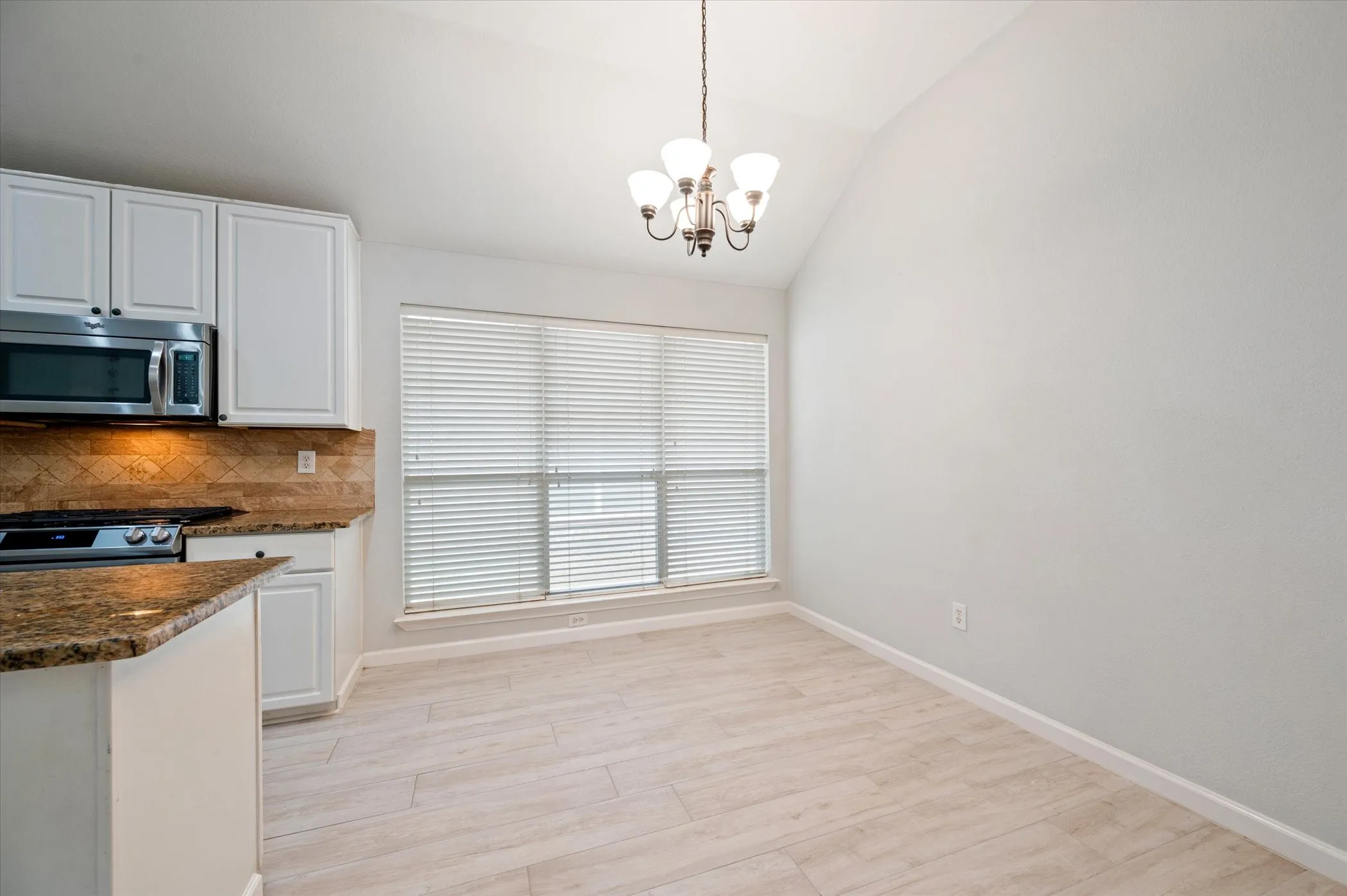 Vaulted ceiling, wood-finish flooring, white cabinetry, integrated stainless steel microwave, and a window with blinds
