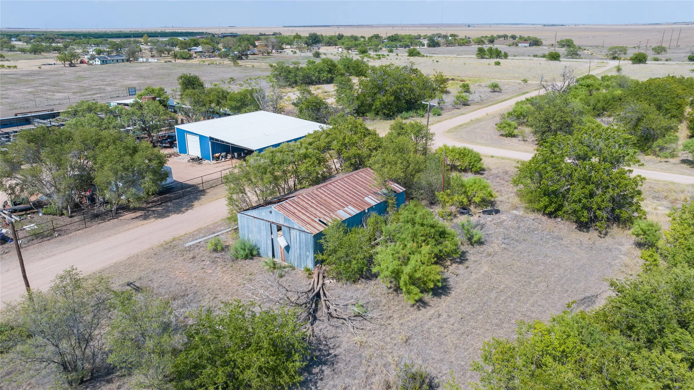 Large blue and white commercial building, secondary metal building with rusted roof, extensive natural landscaping, unpaved roadways, and open terrain