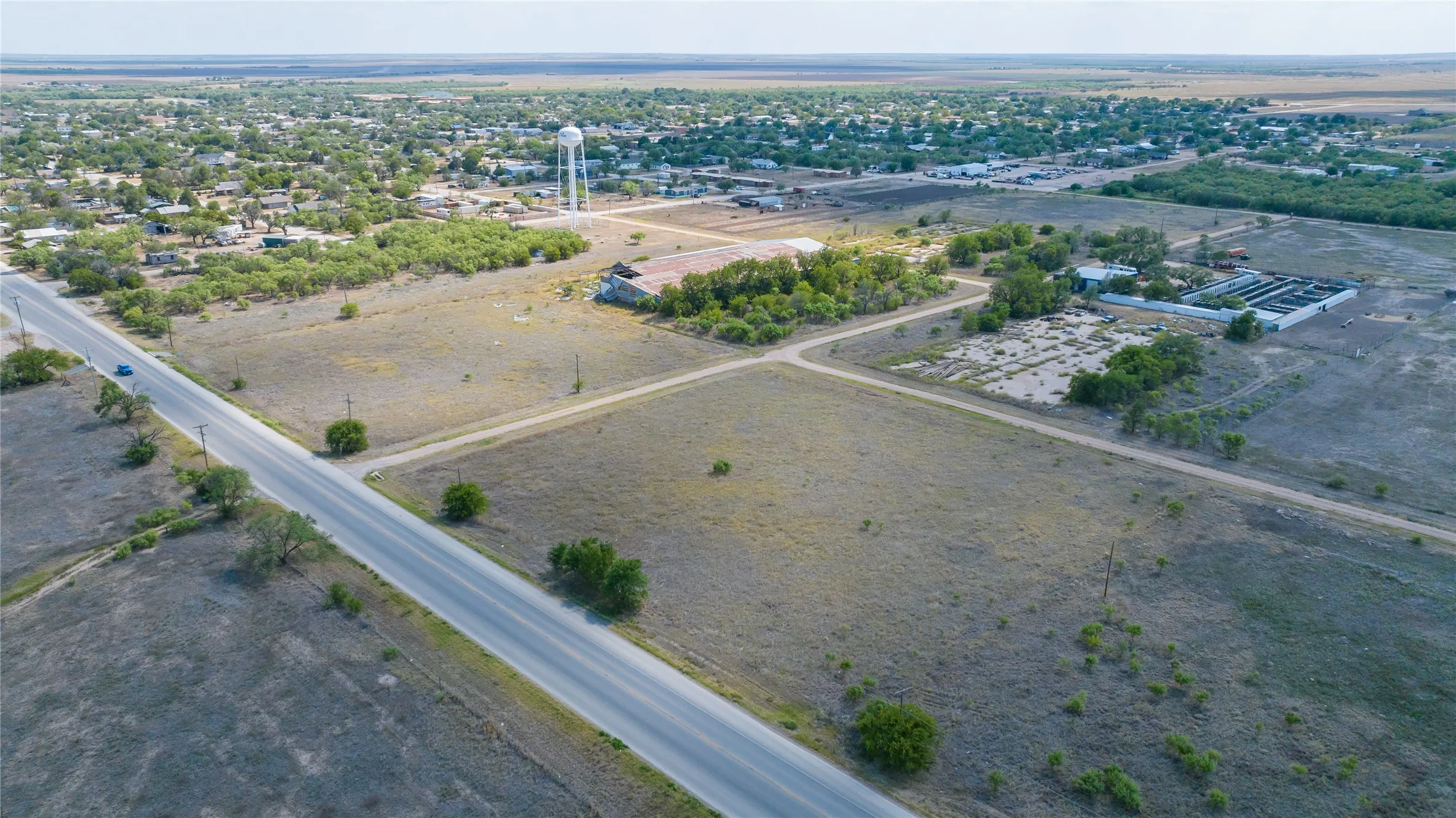 Expansive undeveloped land featuring a paved road, scattered trees, and distant community structures including a water tower