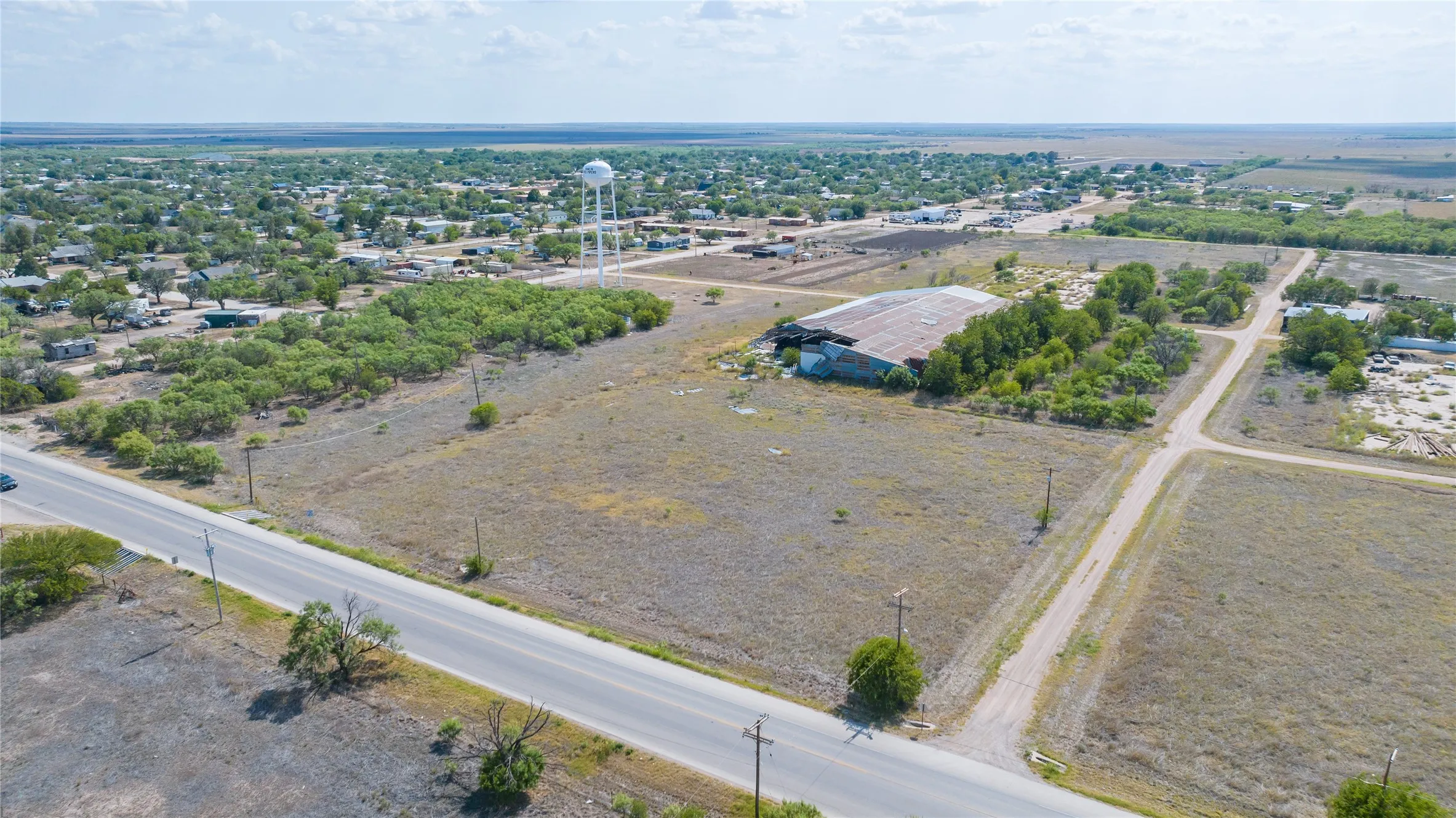 Expansive land parcel featuring paved road frontage, established utility poles, scattered mature trees, and a distant water tower