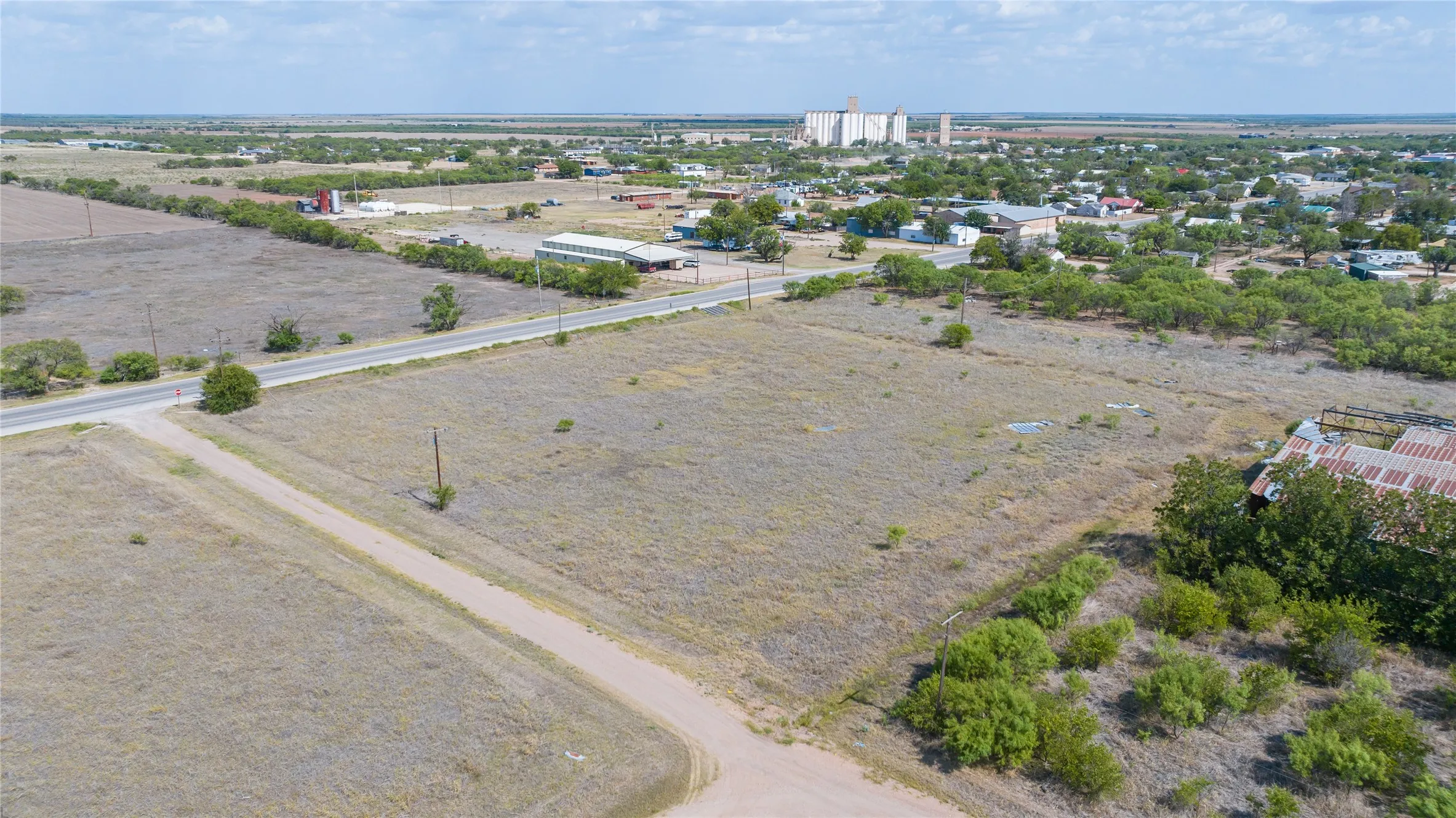 Expansive undeveloped land with a paved road frontage, natural vegetation, and distant town infrastructure