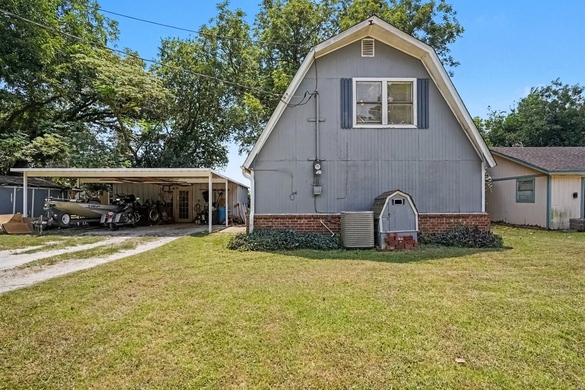 Daytime exterior view displaying the front elevation, attached carport shelter, and mature shade trees.