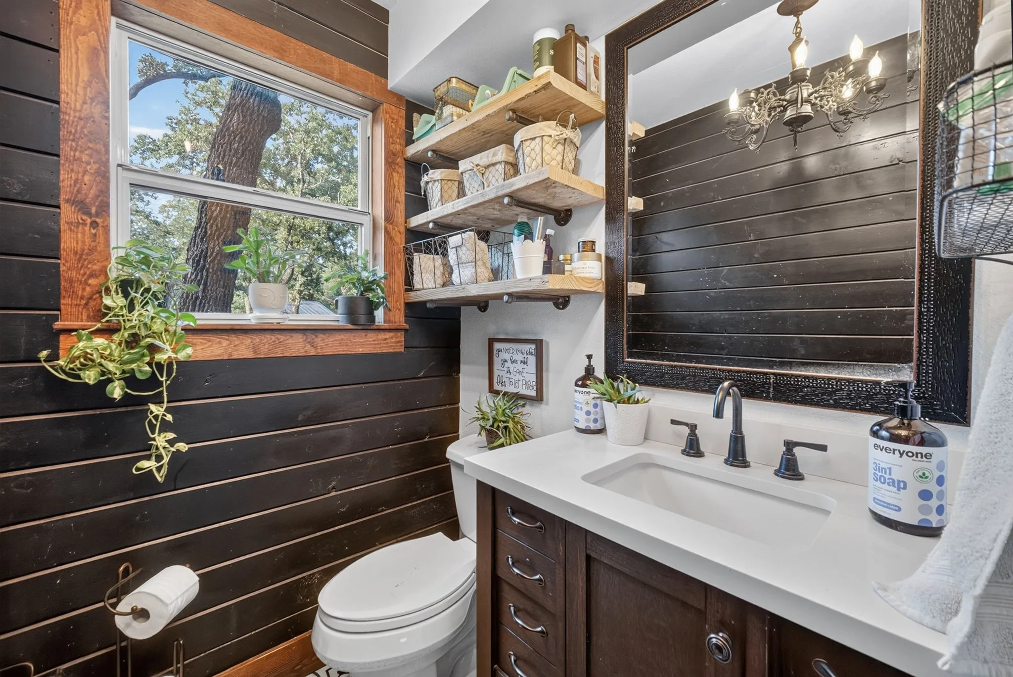 Downstairs powder room. Wood-paneled accent wall with natural wood window trim, white porcelain toilet, wood vanity with white countertop, and mounted shelving