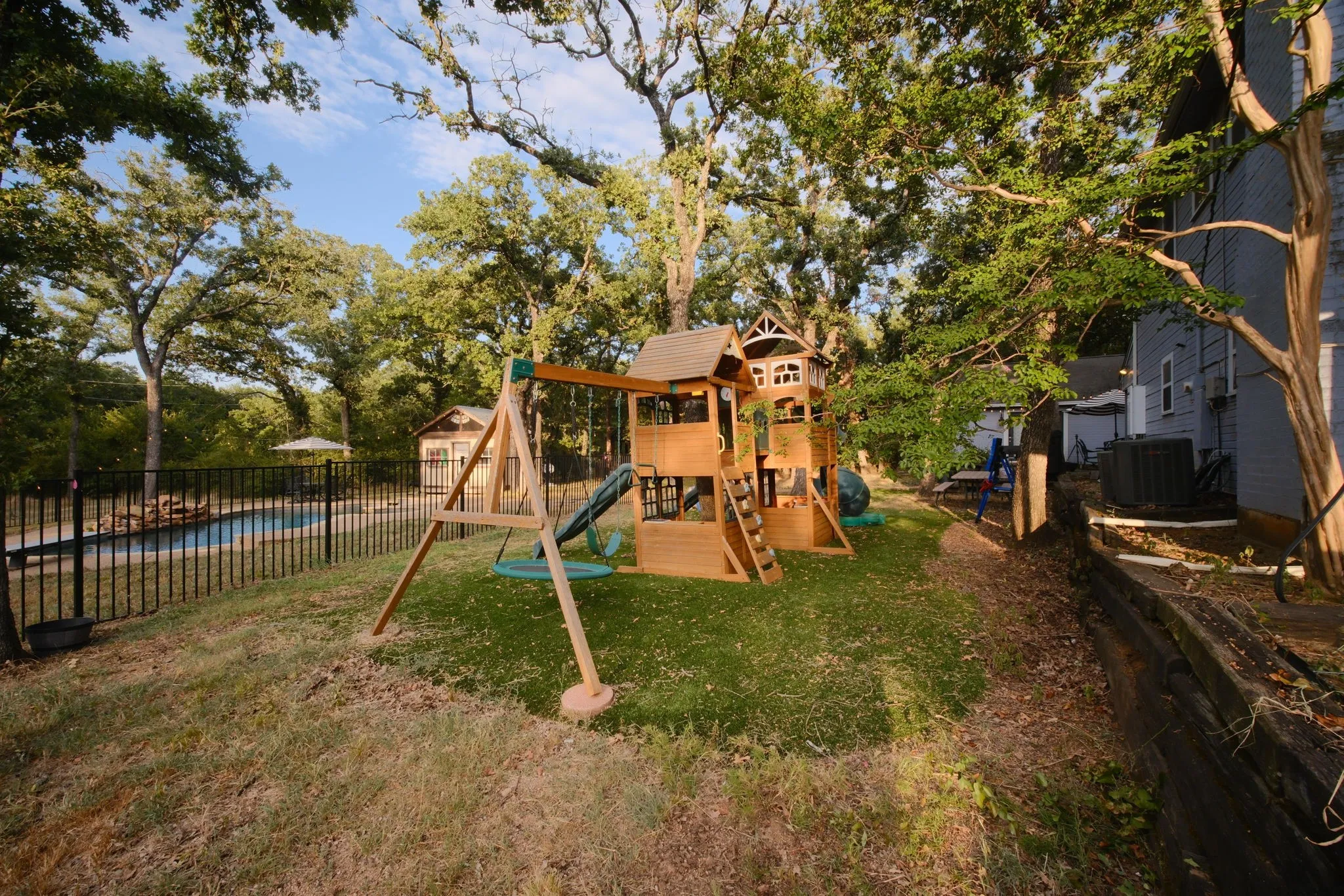 Expansive backyard oasis featuring a multi-level wooden playset with slides and climbing structures, mature shade trees throughout the property, manicured lawn, black iron perimeter fencing, and a detached outbuilding