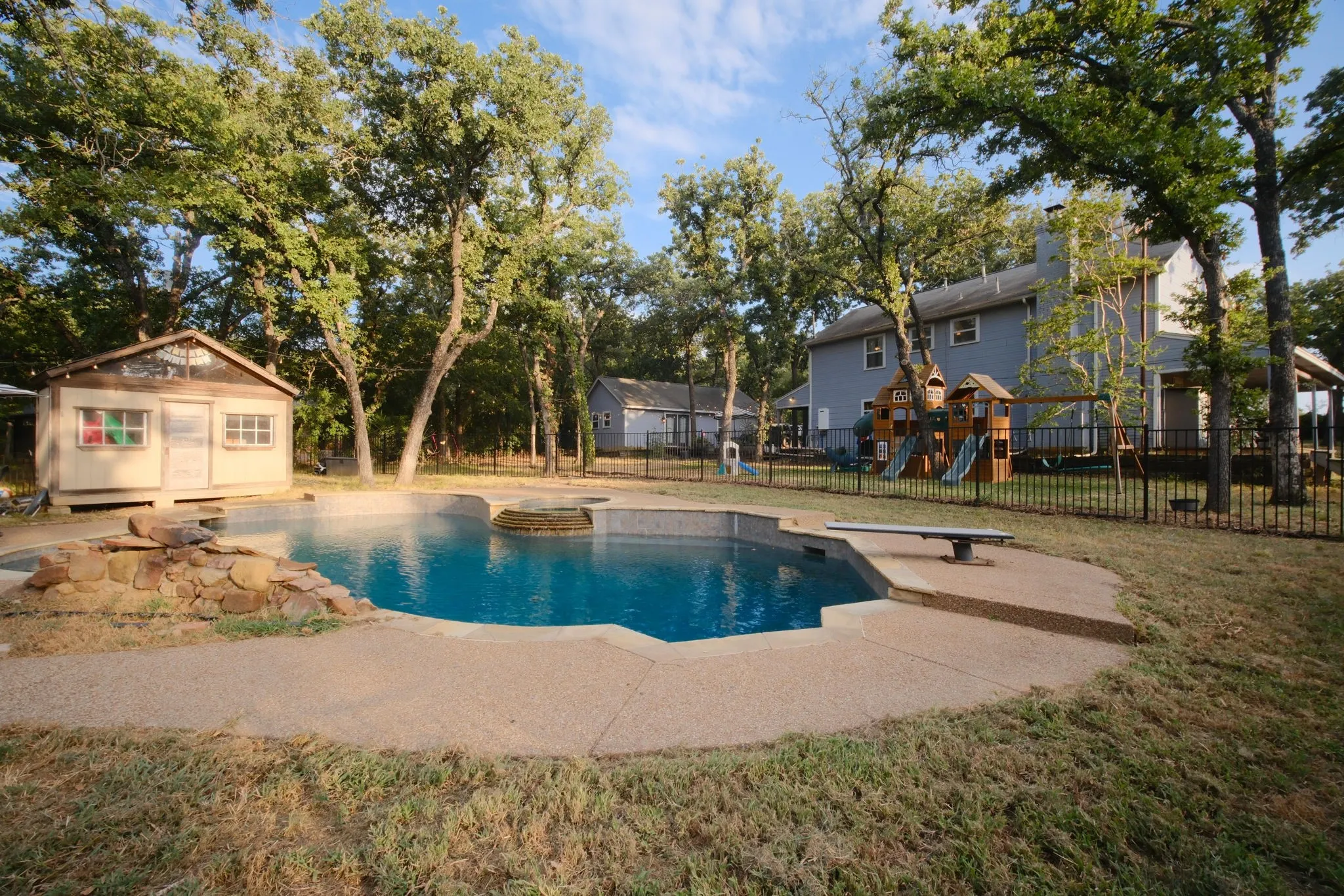 Sparkling swimming pool with sun shelf and integrated spa features, surrounded by a concrete decking and mature shade trees
