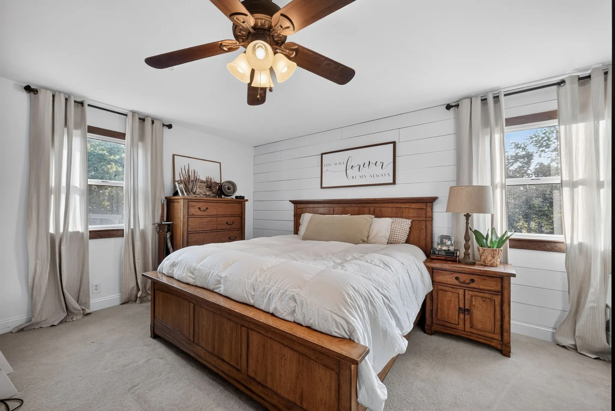 Bright primary bedroom featuring shiplap accent wall, ceiling fan with integrated lighting, dual windows and carpeted flooring throughout