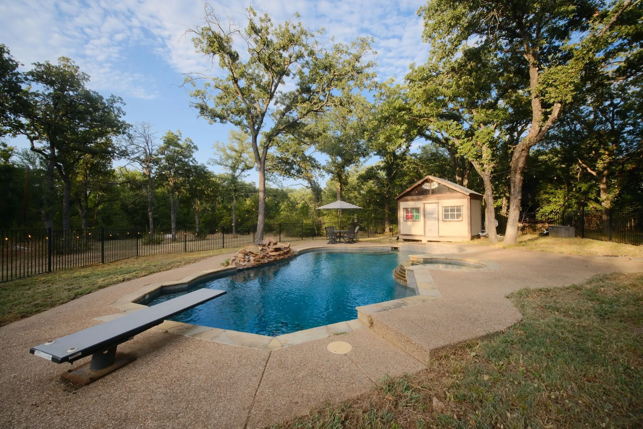 Backyard oasis featuring a sparkling pool with integrated spa, expansive concrete decking, mature shade trees, detached pool house with covered porch, and perimeter fencing