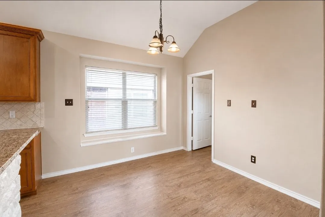 Vaulted ceiling, wood-finish flooring, kitchen cabinetry, tile backsplash, and a window with blinds