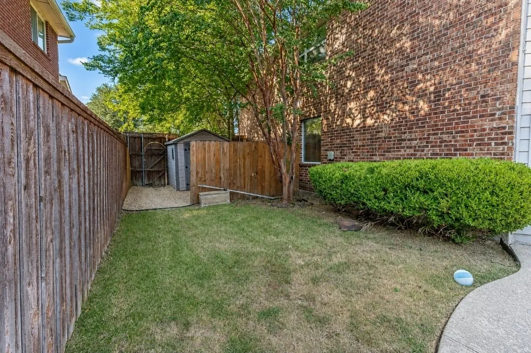 Manicured lawn with mature trees, a privacy fence, and a storage shed on a gravel pad