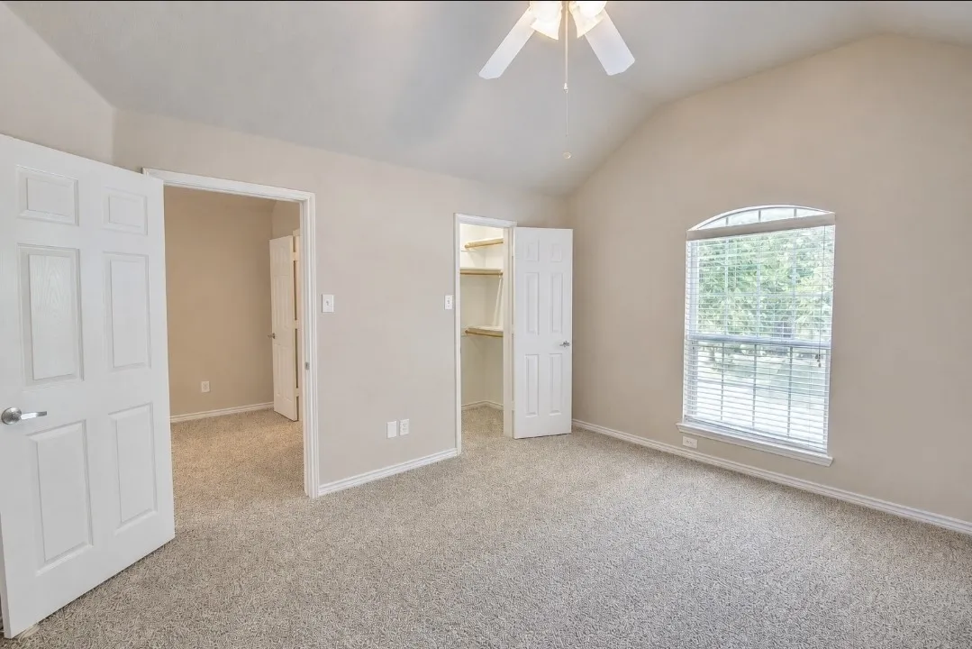Vaulted ceiling with a ceiling fan, arched window with blinds, soft gray carpet flooring, and an open closet with built-in shelving